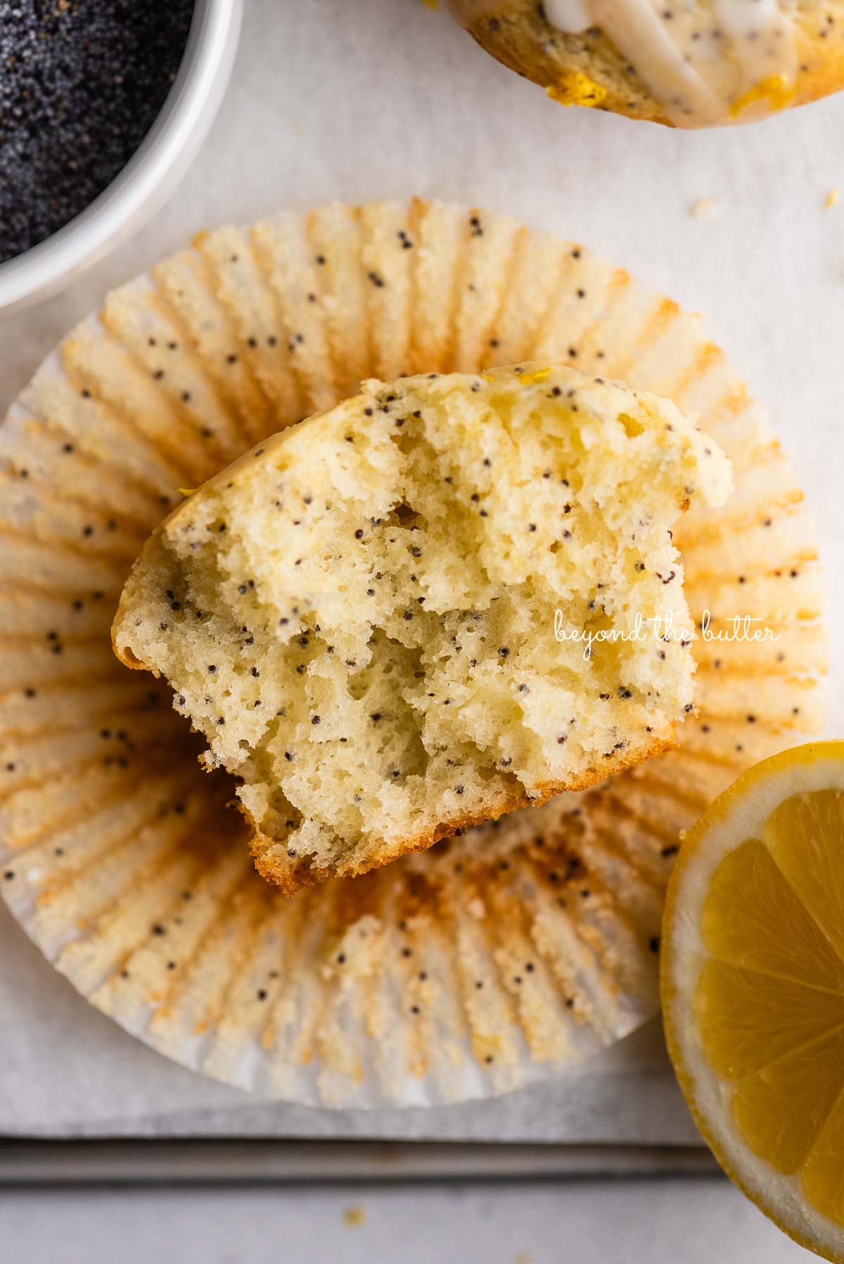 Half eaten lemon poppyseed muffin on top of muffin liner on white background with bowl of poppyseeds and slice of lemon.