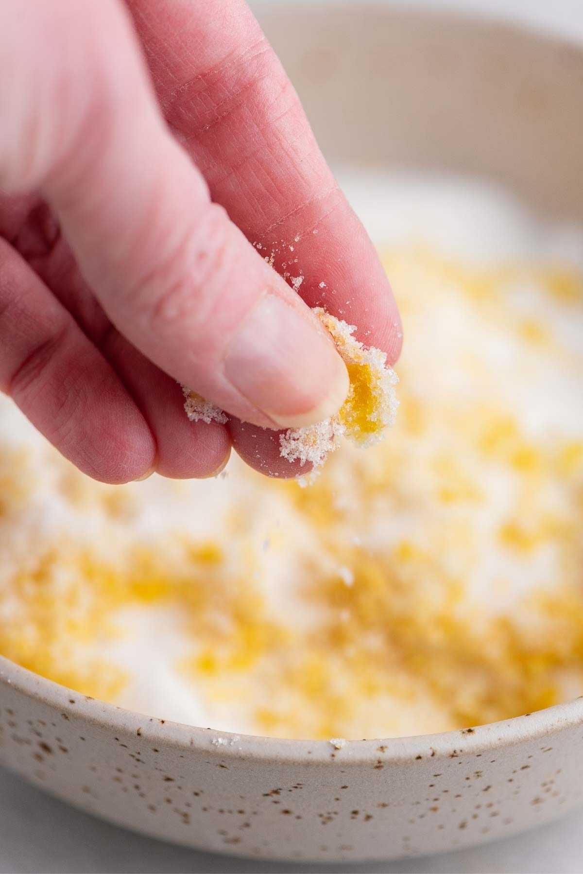 Rubbing sugar and lemon zest together between pointer finger and thumb over a bowl.