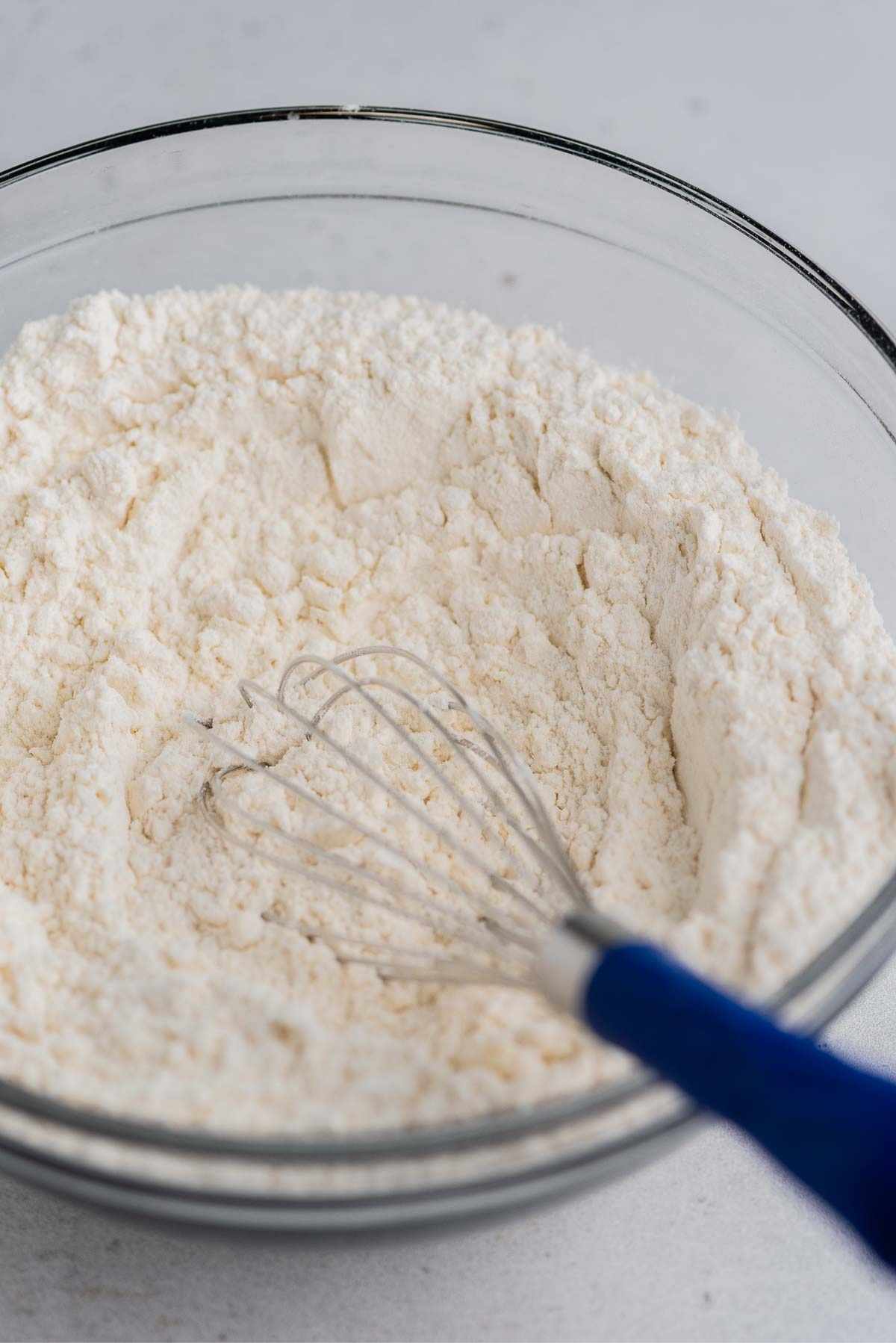 Whisking lemon poppy seed muffin dry ingredients together in a large glass mixing bowl on a light grey background.