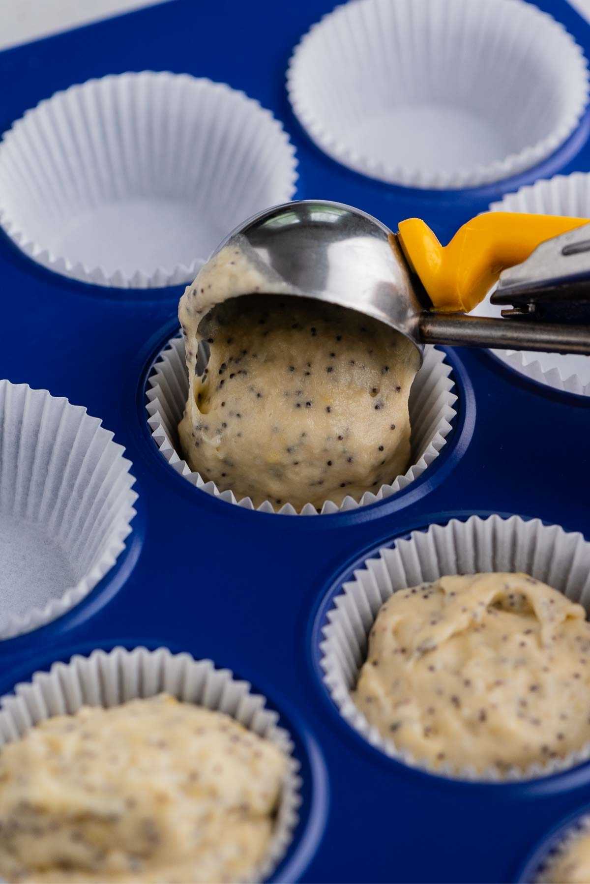 Adding lemon poppy seed muffin batter to a muffin paper liner using a cookie scoop with yellow handle.