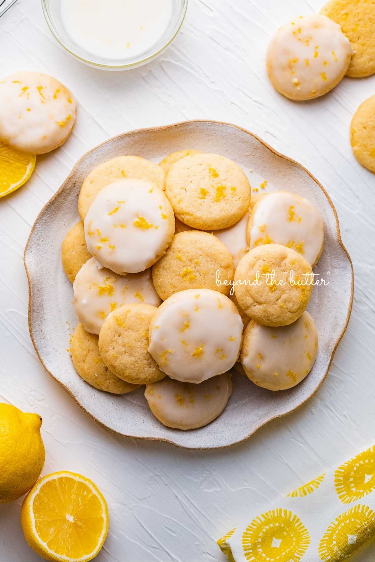 Plate full of lemon shortbread cookies surrounded by a lemon themed cloth napkin, lemons, and glass of milk on a white background.