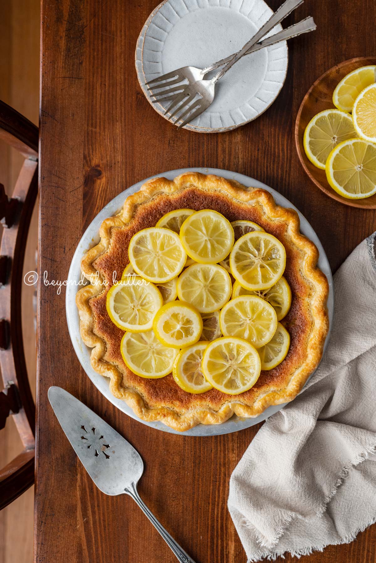 lemon sponge pie decorated with candied lemon sliced on a dark wood background with small dessert plates, napking, pie server, and small plate of sliced lemons | All images © Luyiz®