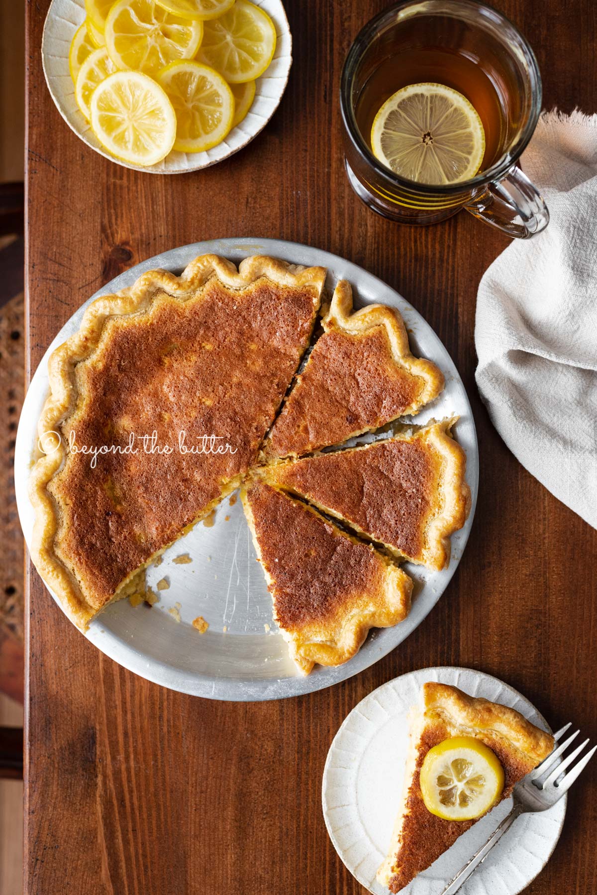 Lemon sponge pie with cut slices on dark wood background with hot lemon tea, small plate of sliced lemons, and slice to the side | All images © Luyiz®