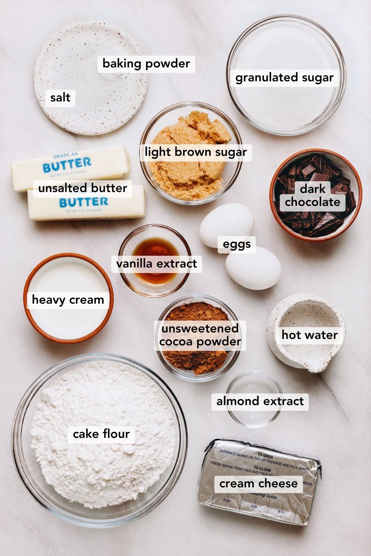 Ingredients in various size bowls and plates labeled as salt, baking powder, granulated sugar, light brown sugar, unsalted butter, heavy cream, vanilla extract, almond extract, cream cheese, hot water, unsweetened cocoa powder, cake flour, dark chocolate, and eggs on a white marbled background.