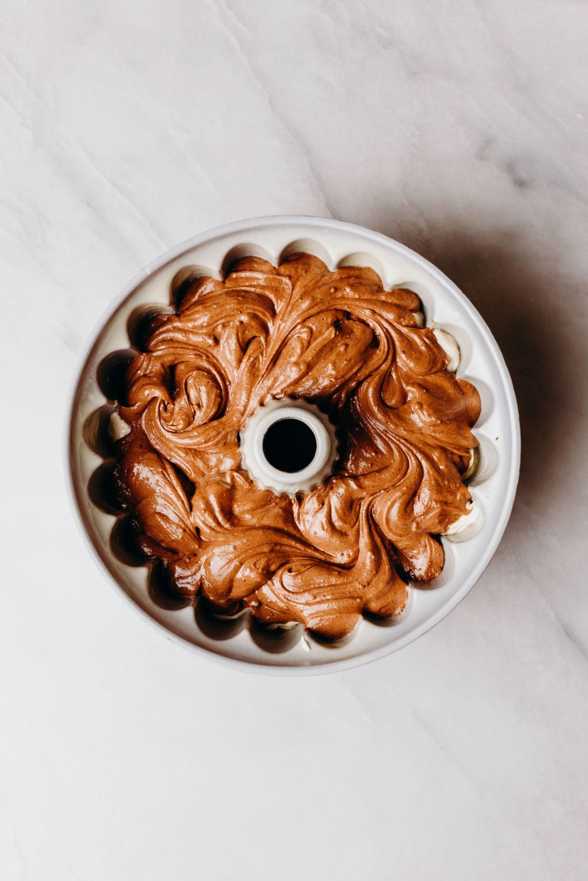 Swirled chocolate and vanilla cake batter in a bundt cake pan on a white marbled background.