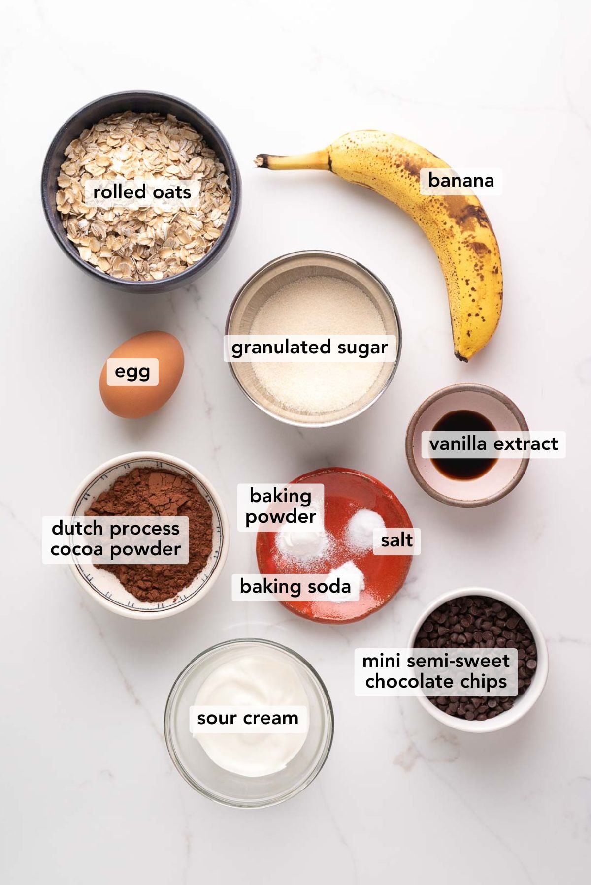 Ingredients in various size bowls labeled as rolled oats, banana, egg, granulated sugar, vanilla extract, dutch process cocoa powder, baking powder, salt, baking soda, mini semi-sweet chocolate chips, and sour cream on a white marbled background.