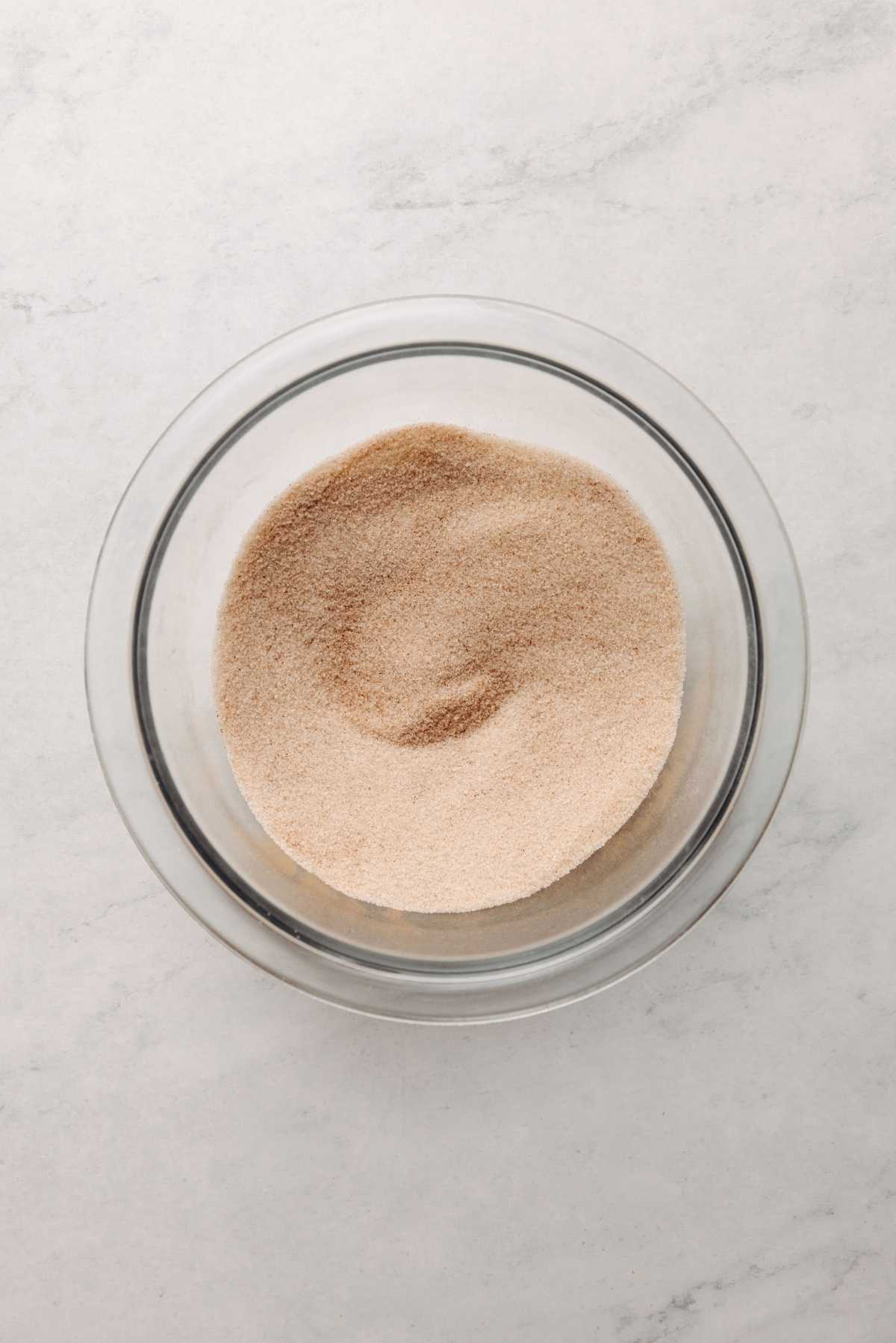 Cinnamon and sugar whisked together in small glass mixing bowl on a white marbled background.