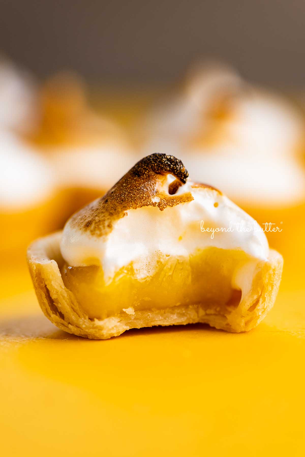 Mini lemon meringue pie with bite taken out surrounded by other mini pies on a yellow background | © Luyiz@