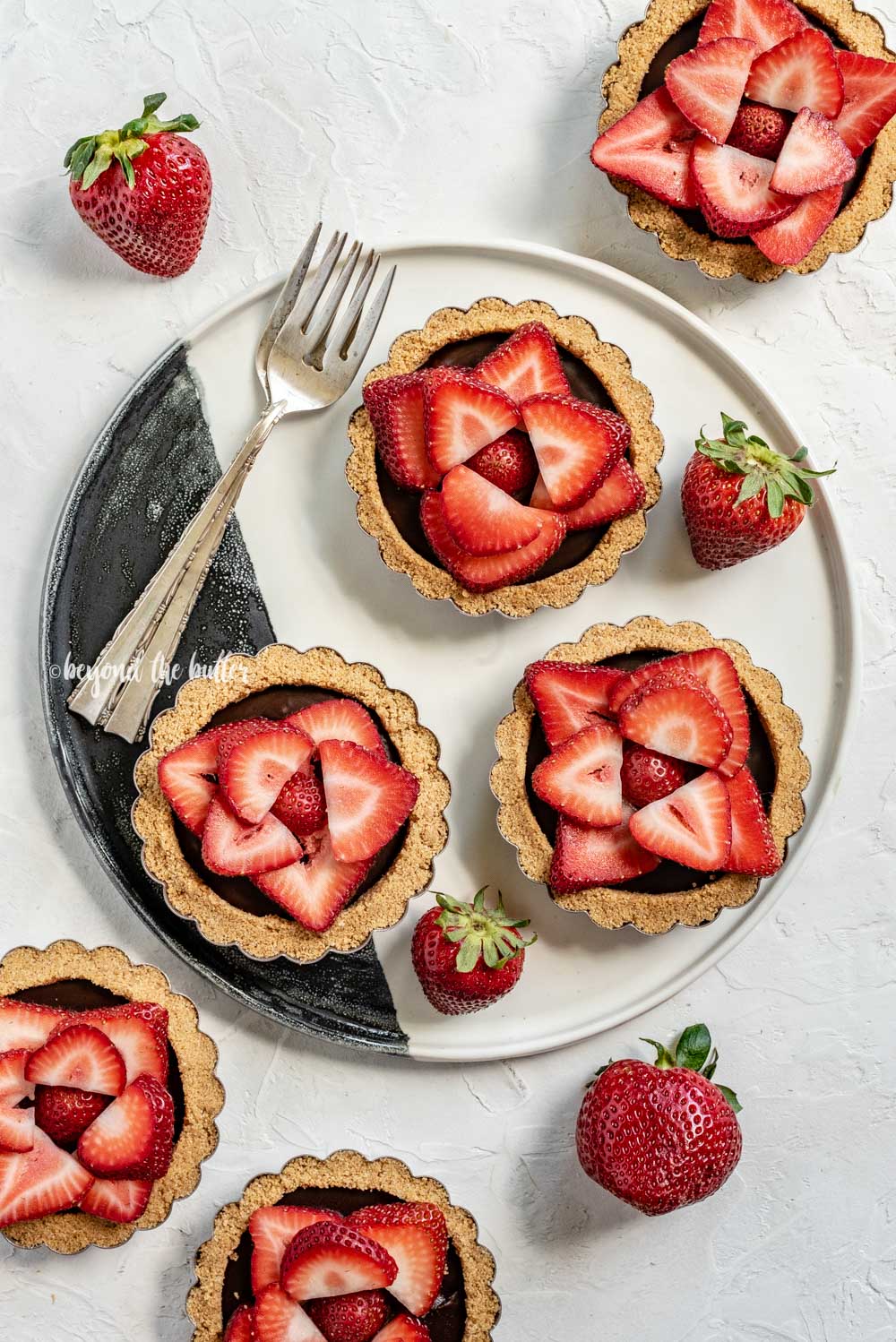 Overhead image of 6 mini strawberry nutella tarts on a black and white serving plate and randomly placed strawberries | All Images © Luyiz™