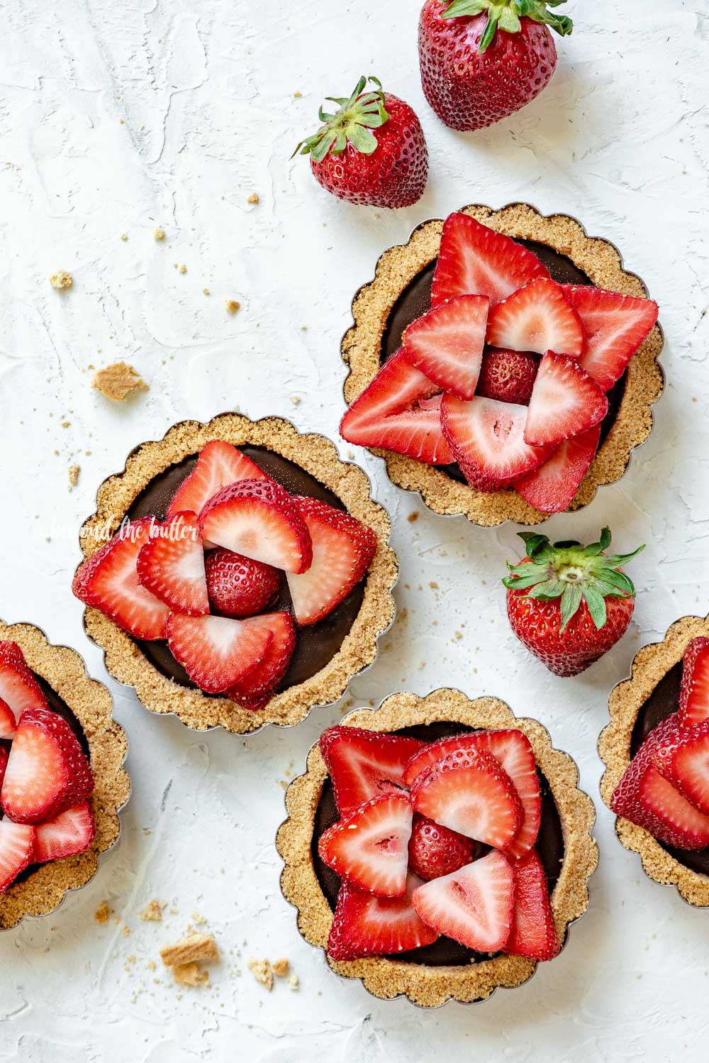 Overhead image of 6 mini strawberry nutella tarts on white stucco background with randomly placed strawberries around them | All Images © Luyiz™