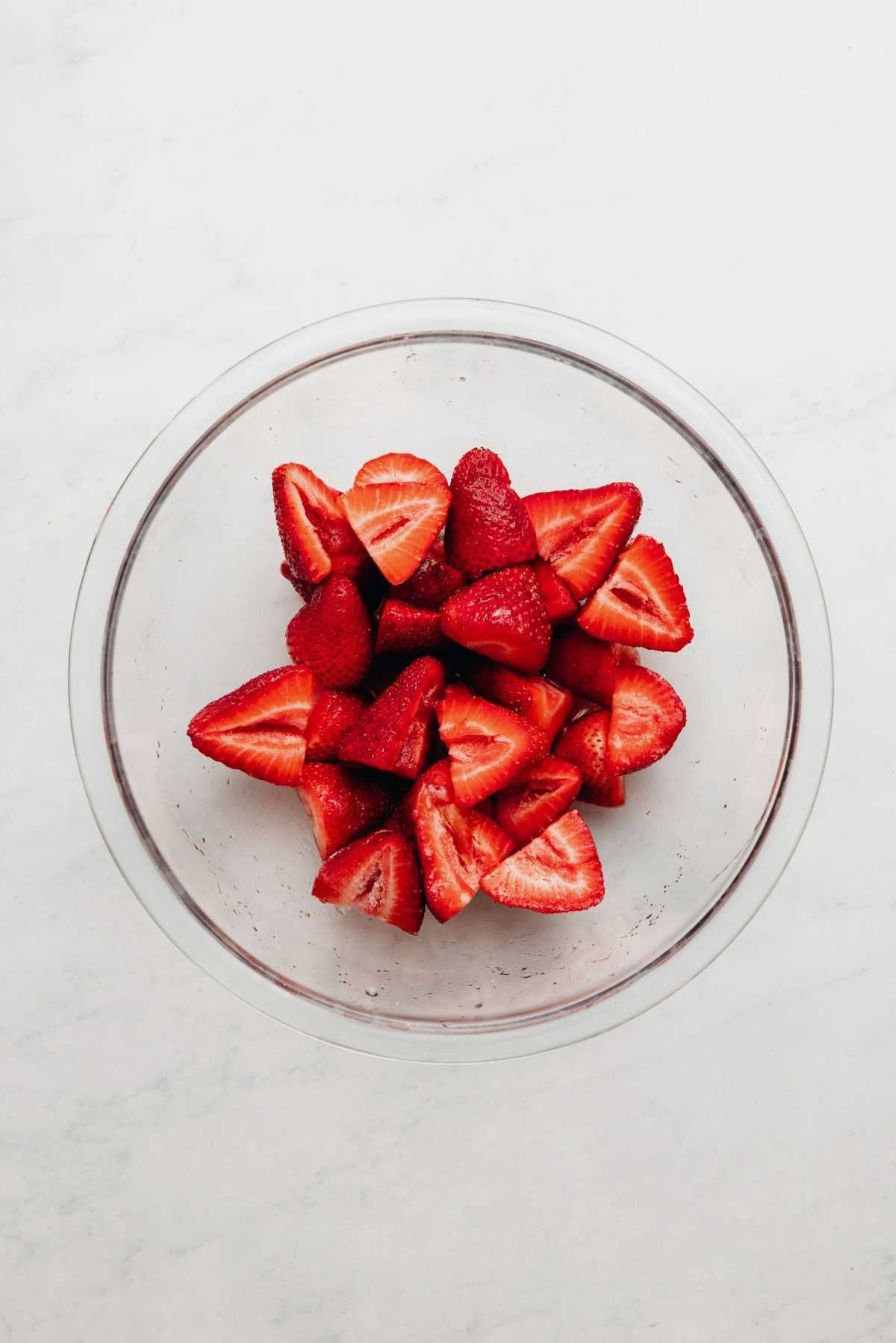 Sliced strawberries, lemon juice, and granulated sugar tossed together in a small glass bowl on a white and grey textured background.
