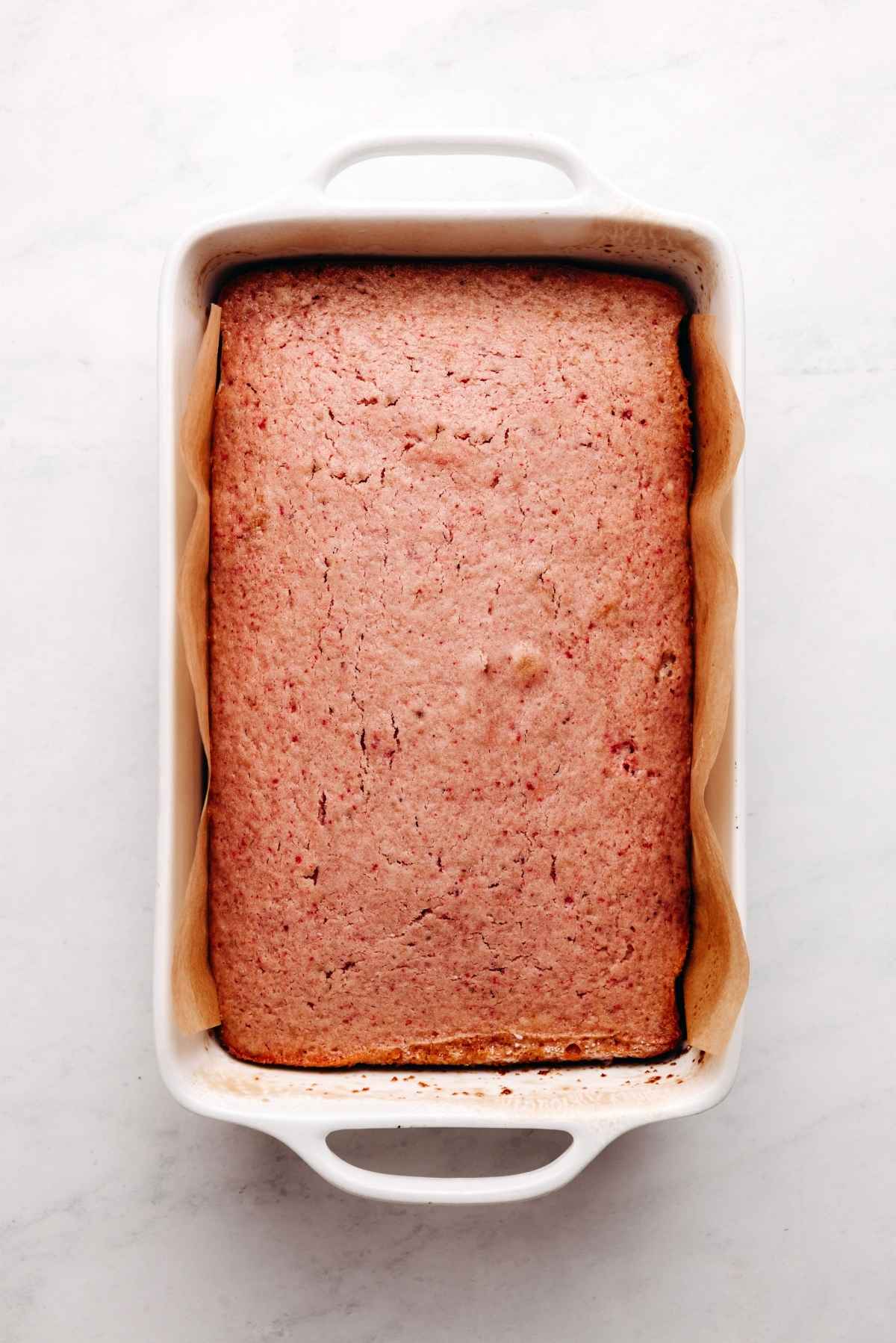 Baked mini strawberry sheet cake in a parchment paper lined baking tin on a white and grey textured background.