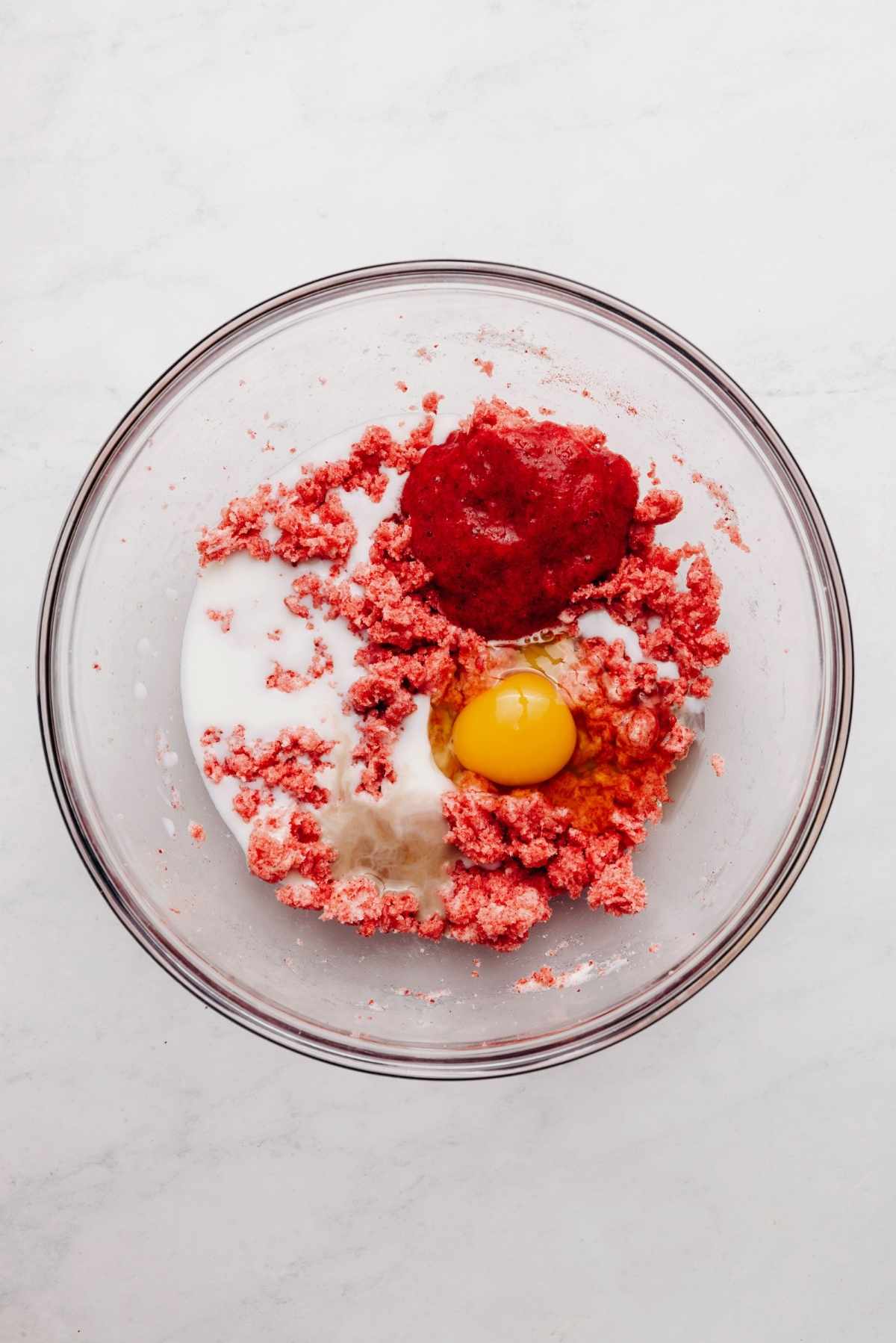 Vanilla extract, milk, roasted strawberry puree, and an egg added to the top of the cake mixture in a glass mixing bowl.