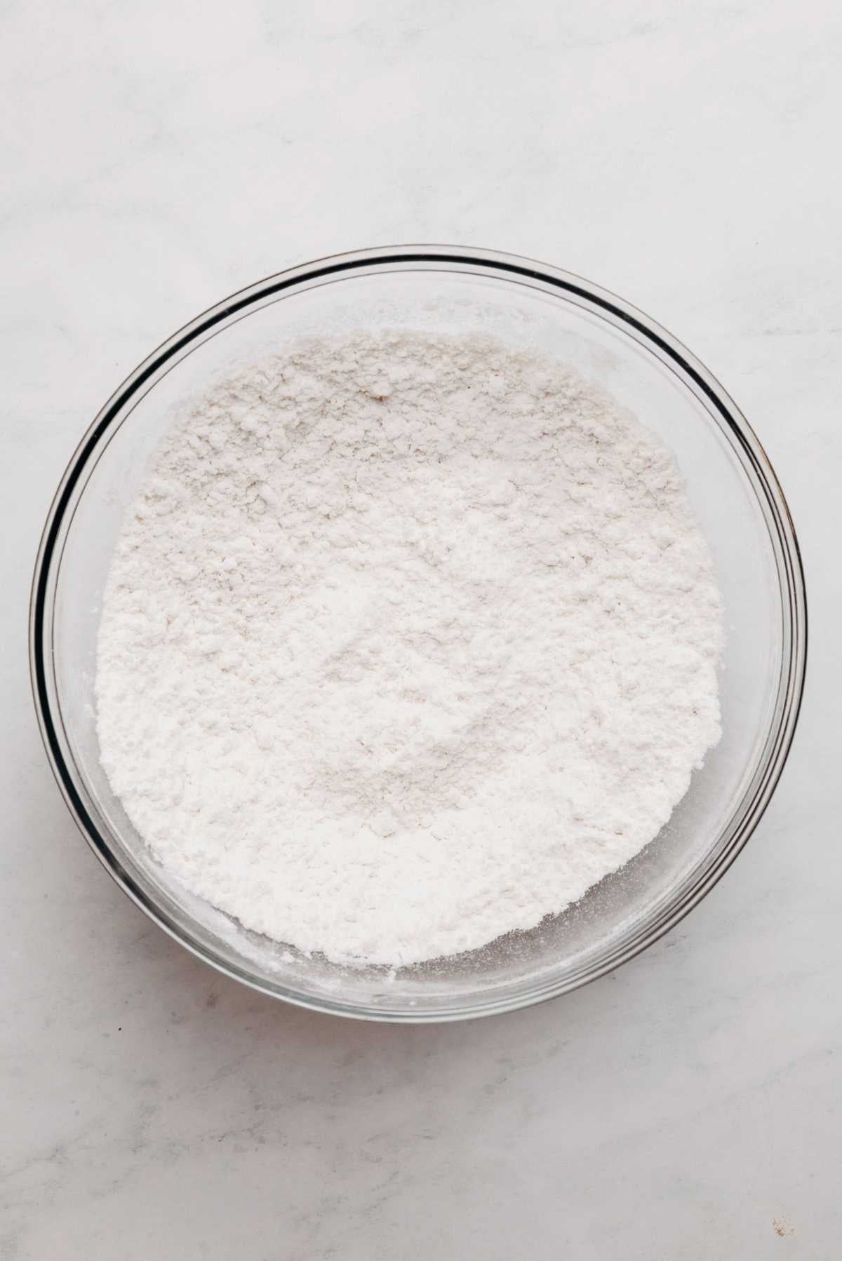 Whisked cake flour, baking powder, and salt in large glass mixing bowl on a white and grey textured background.