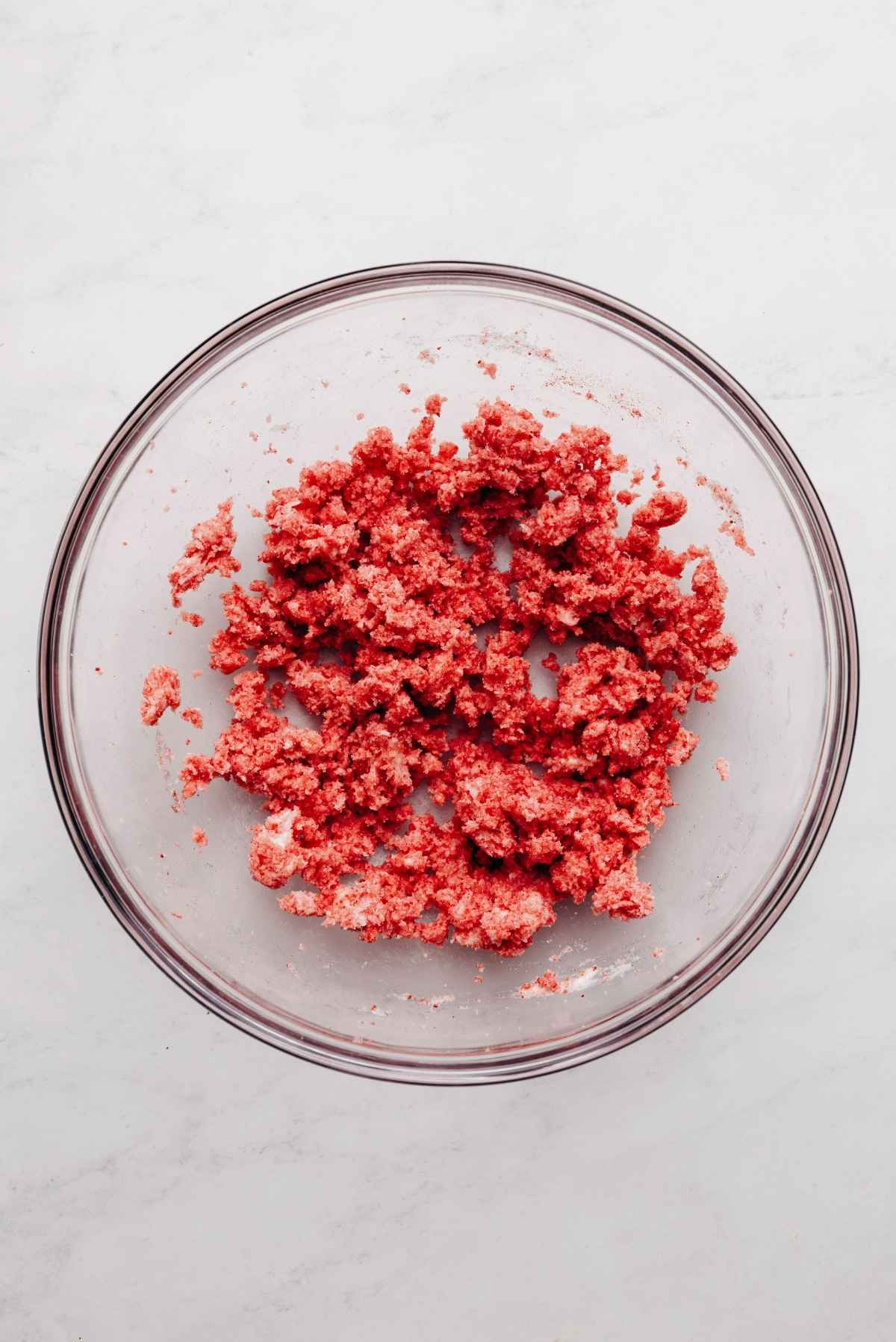 Freeze dried strawberry powder, butter, and sugar blended together in a glass mixing bowl on a white and grey textured background.