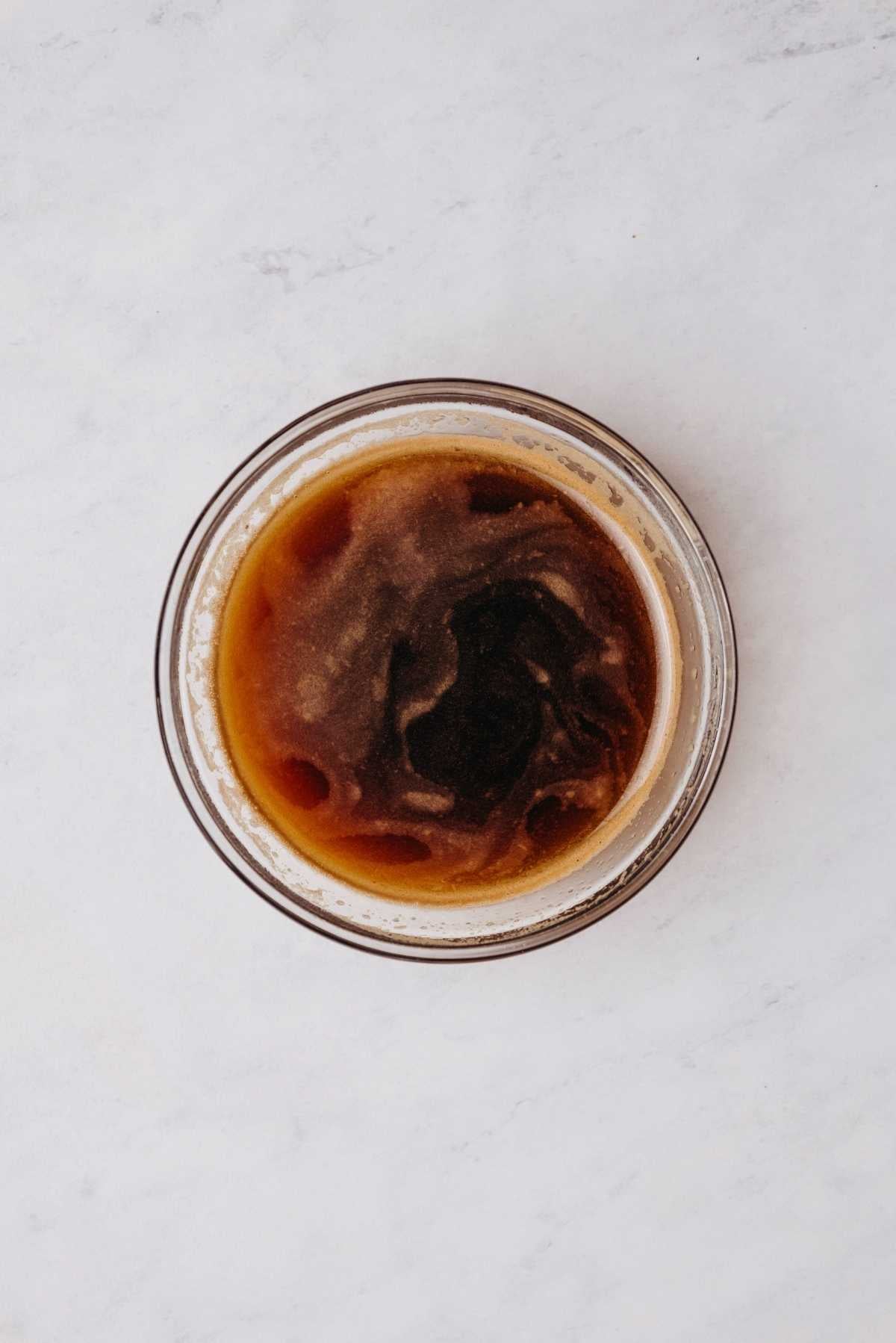 Espresso powder and hot water whisked together in a small glass mixing bowl on a grey marbled background.