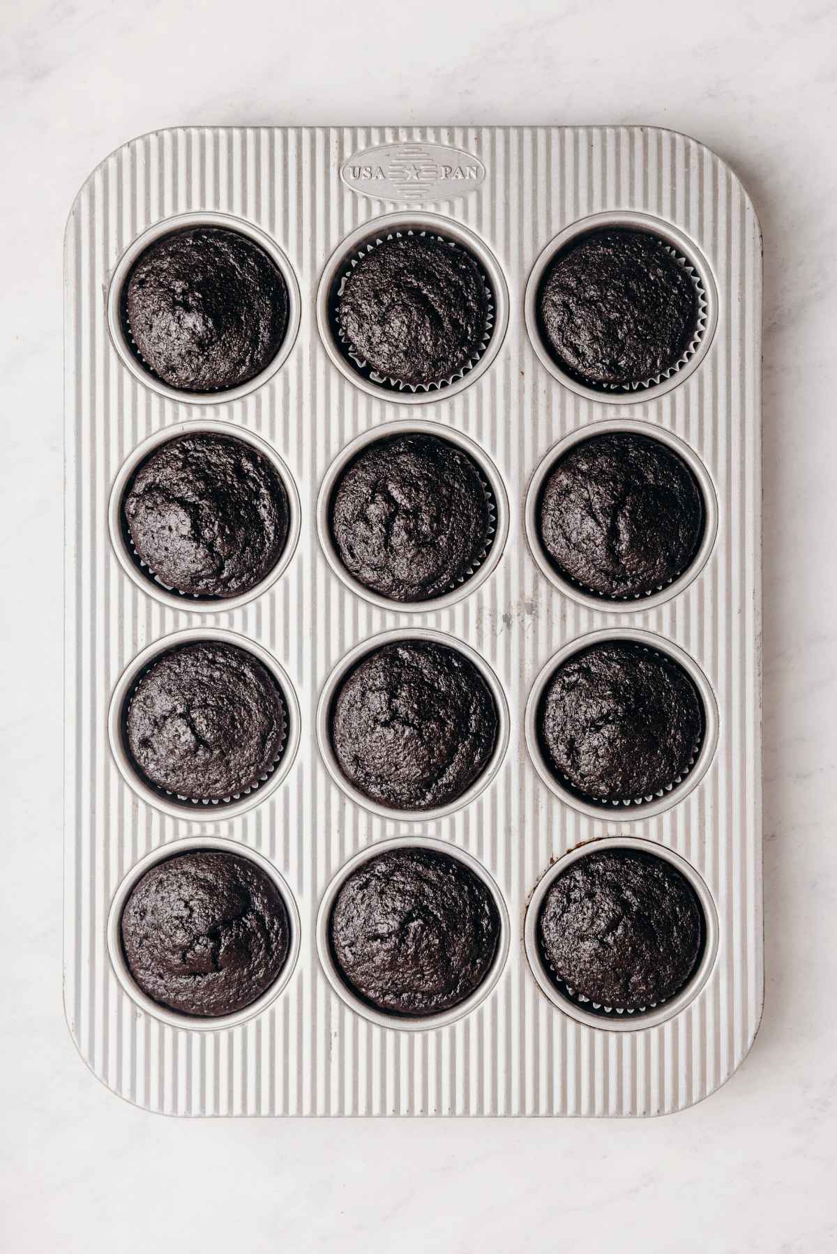 Baked chocolate cupcakes with cupcake liners in a metal cupcake tin on a grey marbled background.