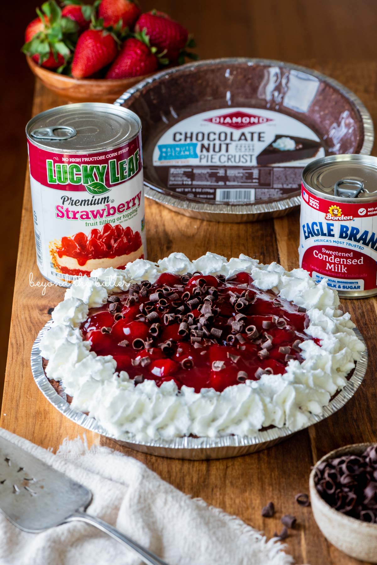 Diamond of CA® chocolate nut pie crust, Lucky Leaf® strawberry pie filling, and Eagle Brand® sweetened condensed milk with decorated no bake strawberry chocolate pie with small bowl of chocolate curls and strawberries on wood table | All images © Luyiz®