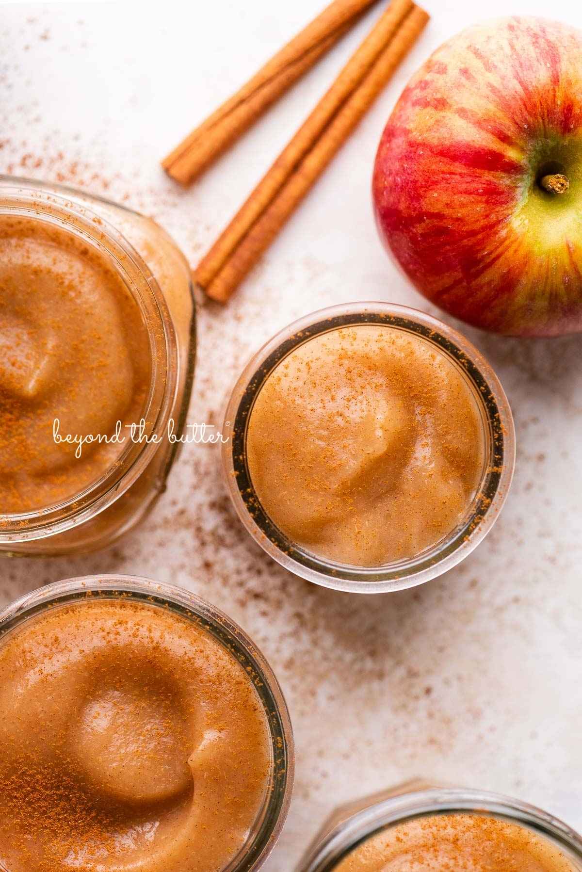 Jars of no sugar added applesauce with cinnamon sticks and apples on a white background.