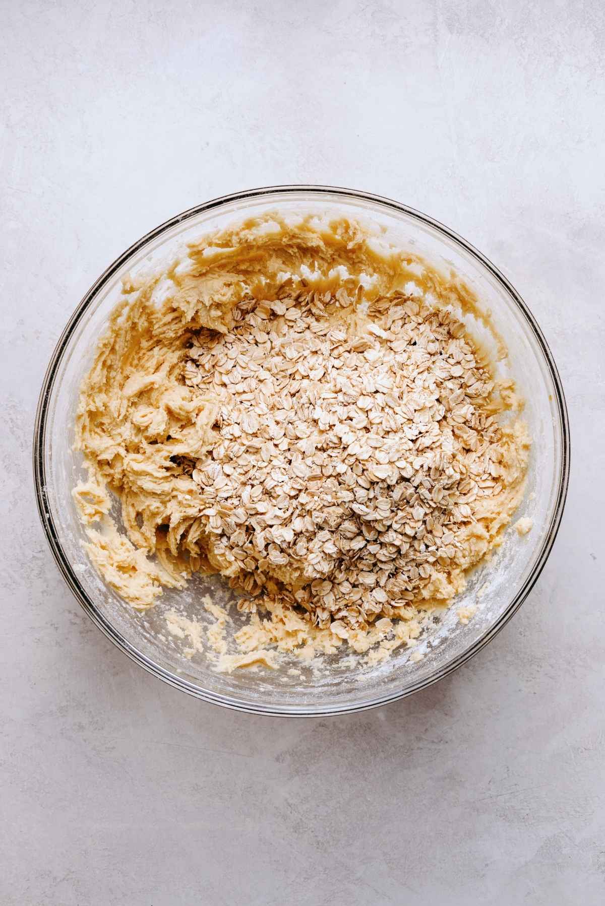 Rolled oats added to the top of oatmeal cookie dough mixture in a glass mixing bowl on a white grey marbled background.
