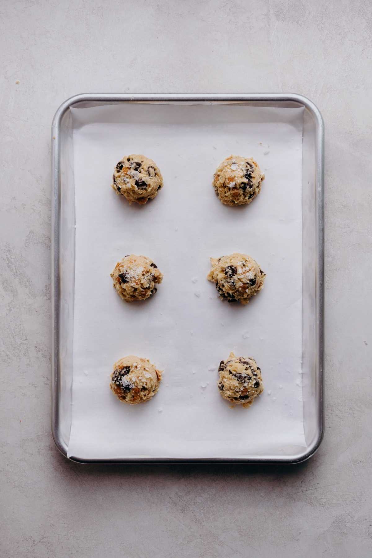 Scoops of oatmeal chocolate chip salted caramel cookie dough topped with flaky sea salt on a white parchment paper lined cookie sheet.