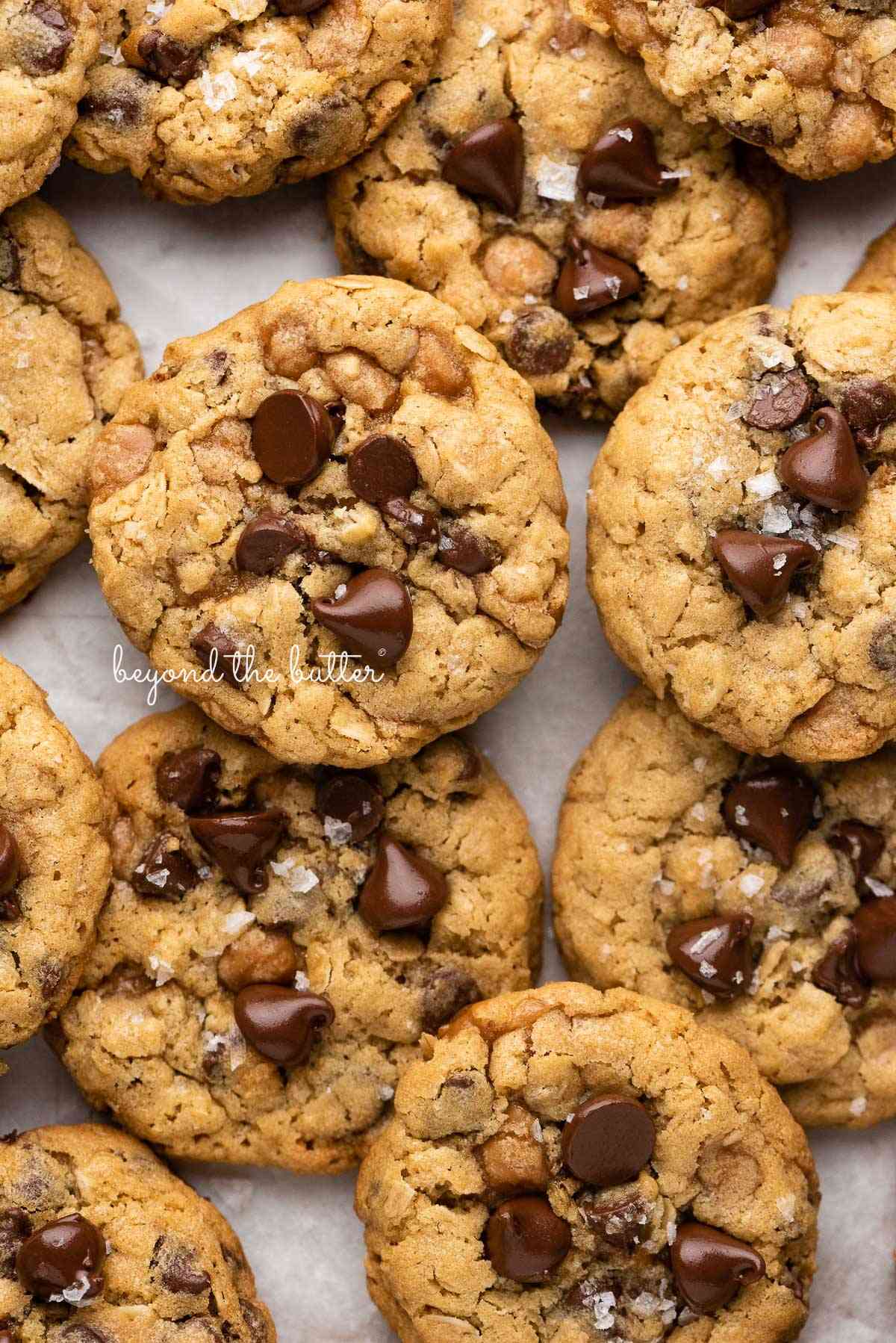 Oatmeal chocolate chip salted caramel cookies topped with flaky sea salt on a parchment paper lined baking sheet.
