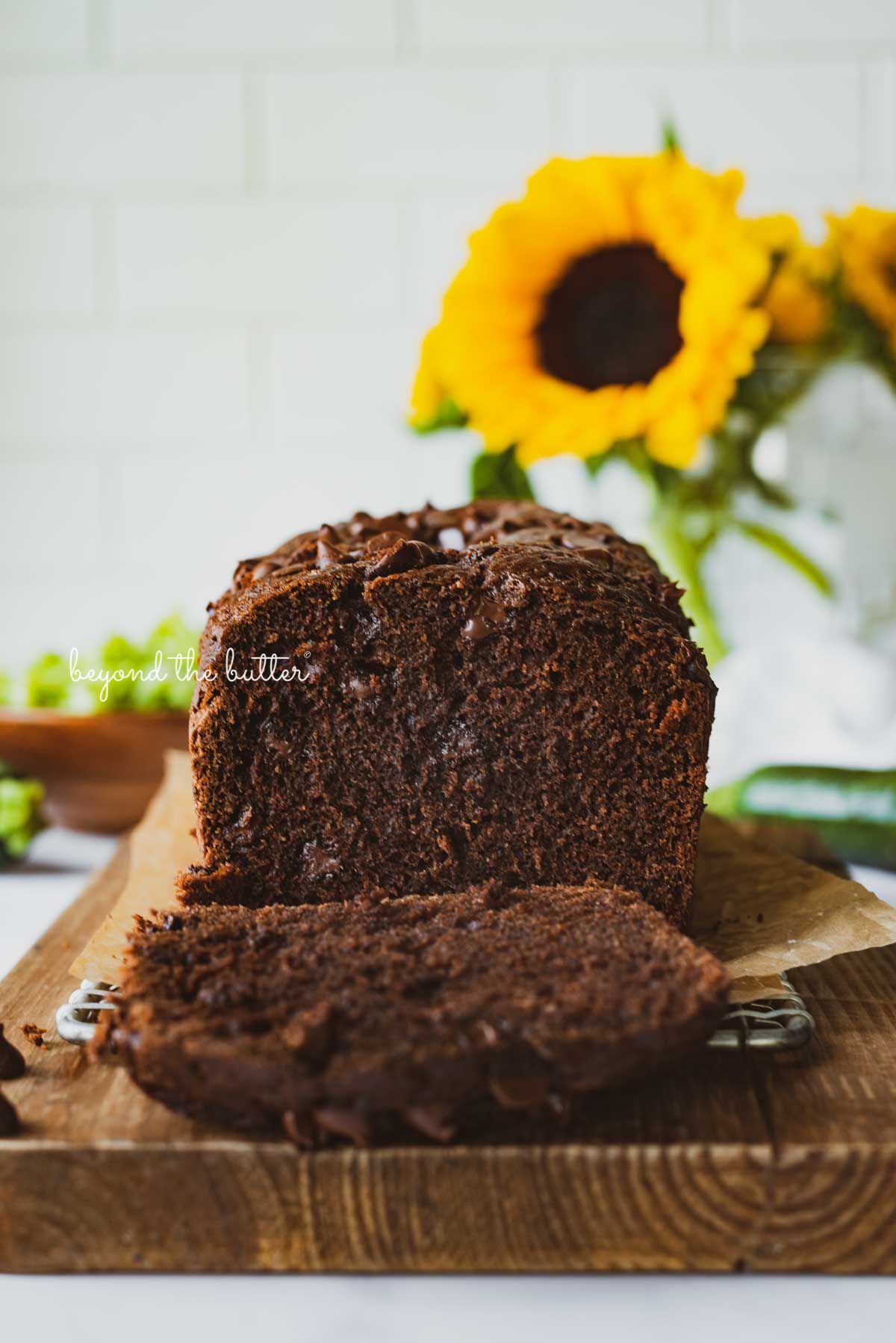 Sliced chocolate zucchini bread on a cutting board with white tile background and sunflowers | © Luyiz®