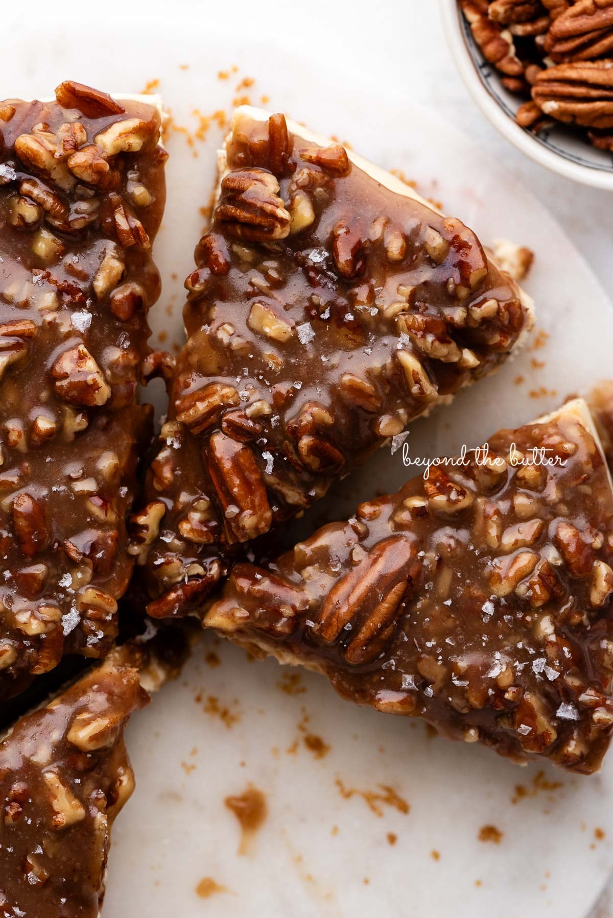Slices of pecan pie cheesecake topped with sea salt flakes on a white marble cake stand next to a small bowl of pecans on a white background.