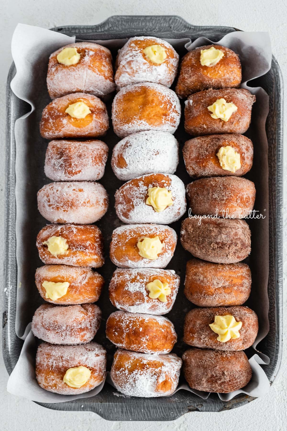 Rows of pennsylvania dutch fasnacht doughnuts in a parchment paper lined baking tray filled with pastry cream | © Luyiz®