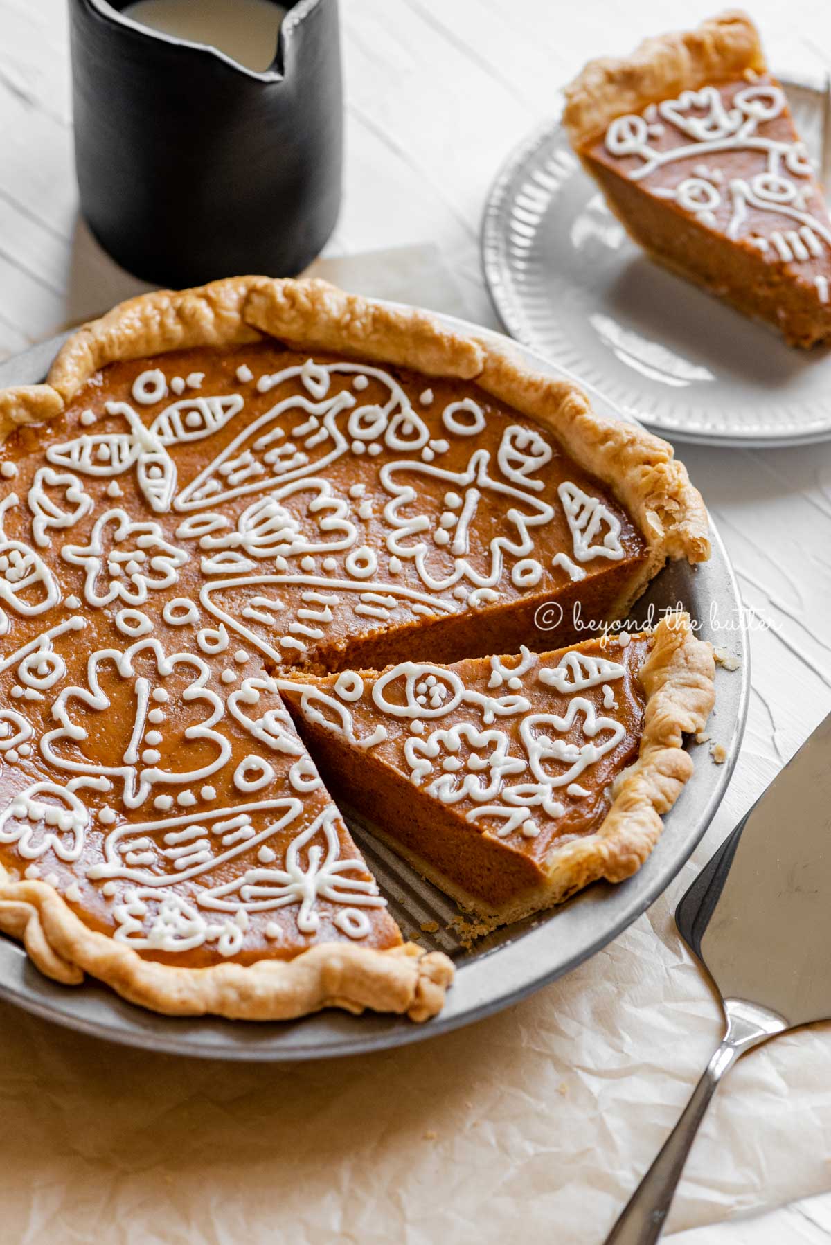 Angled image of cut pumpkin pie with a piped cream cheese frosting decoration surrounded by pie server, cinnamon sticks, plates, and small pitcher of milk | All images © Luyiz™