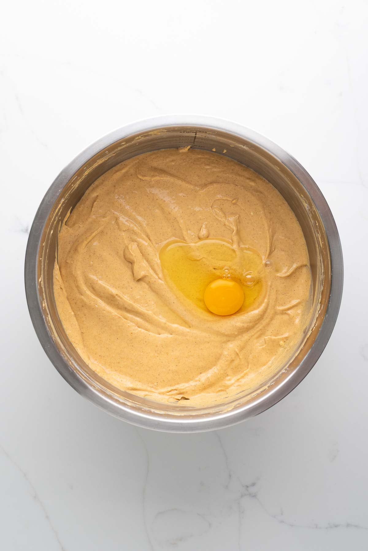 Egg added to pumpkin cheesecake batter in a stainless steel mixing bowl on a white marbled background.