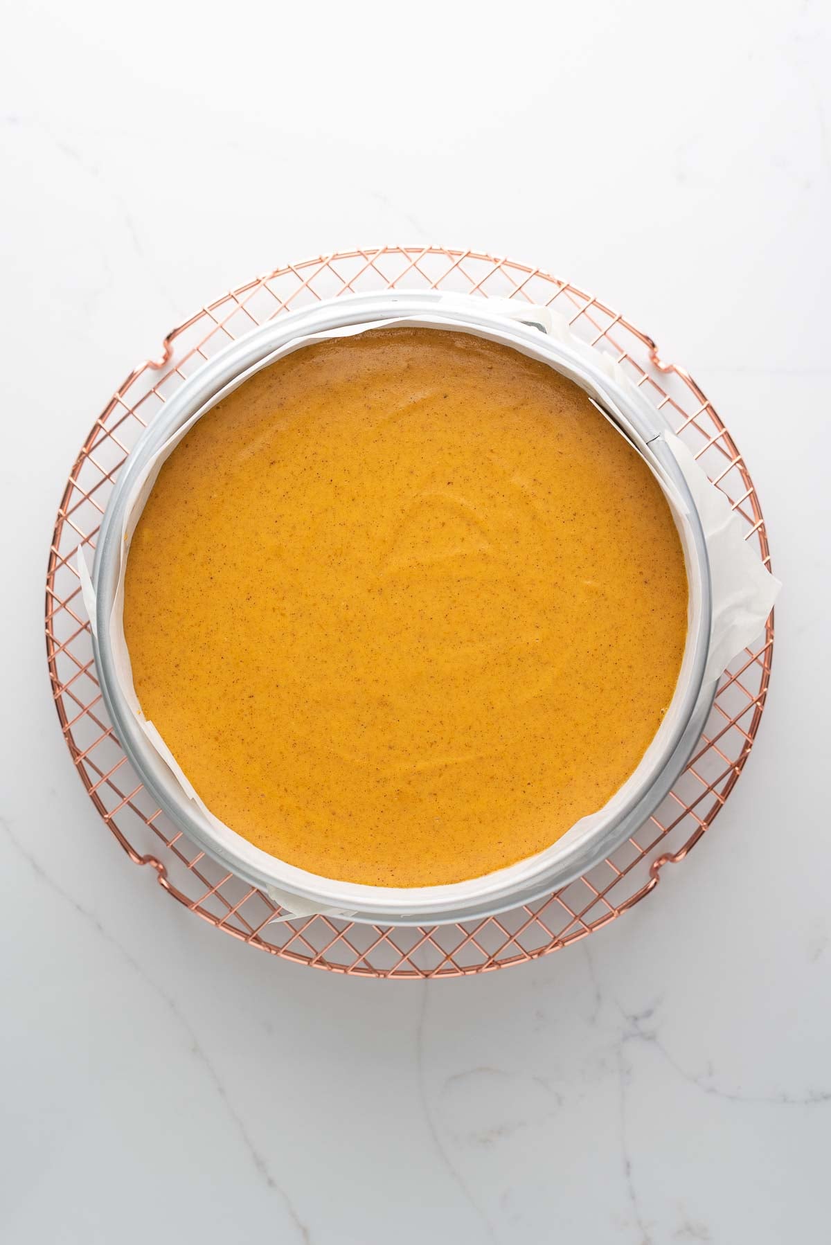 Cooling a pumpkin cheesecake on a round rose gold colored cooling rack on a white marbled background.