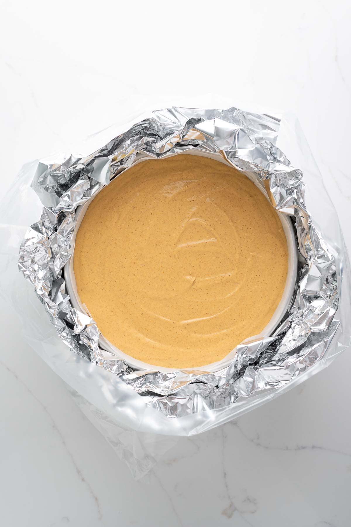Pumpkin cheesecake in a parchment paper lined springform pan wrapped in aluminum foil and a slow cooker bag on a white marbled background.