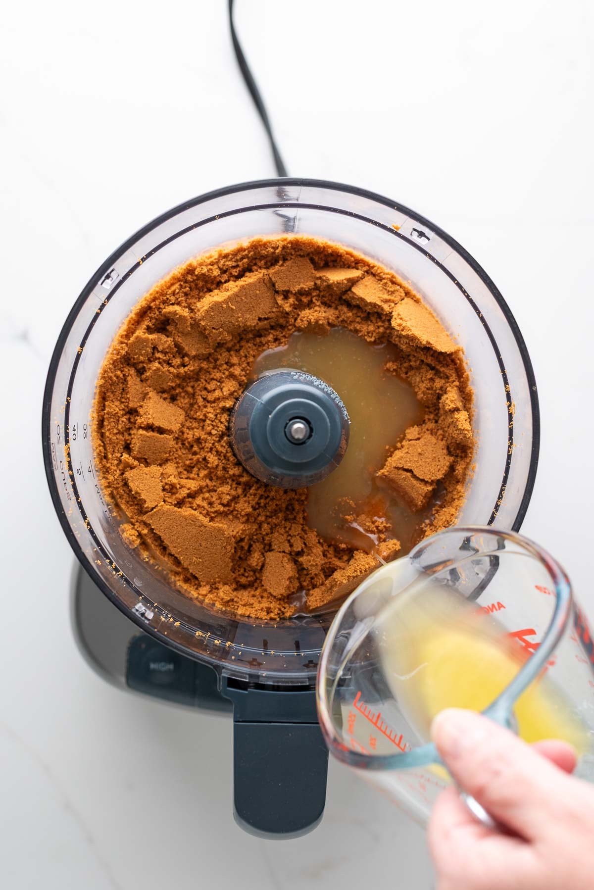 Adding melted unsalted butter in a glass measuring cup to a food processor with speculoos cookie crumbs.