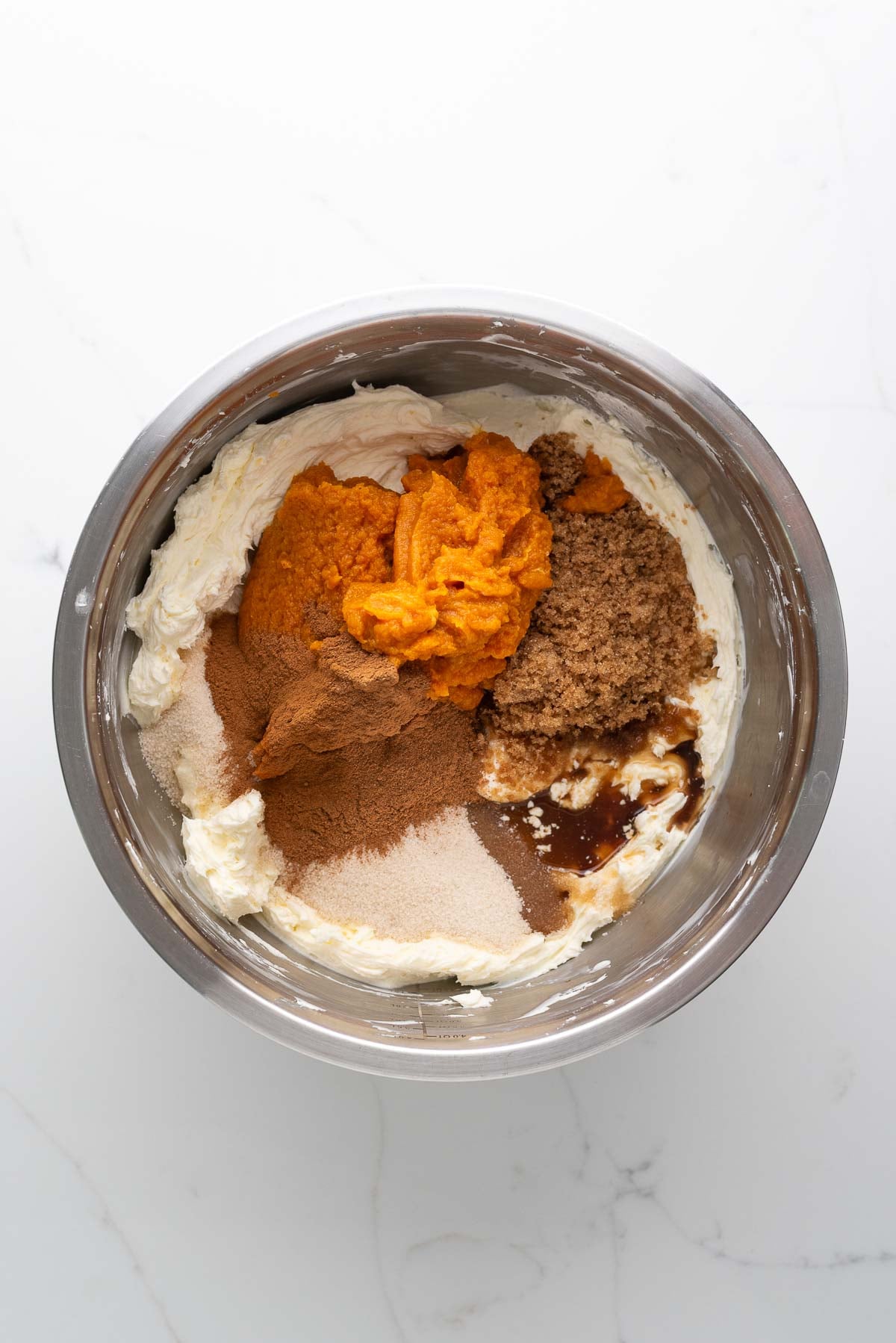 Pumpkin puree, granulated sugar, light brown sugar, vanilla extract, cinnamon, sour cream, and pumpkin pie spice added to the whipped cream in a stainless steel mixing bowl.