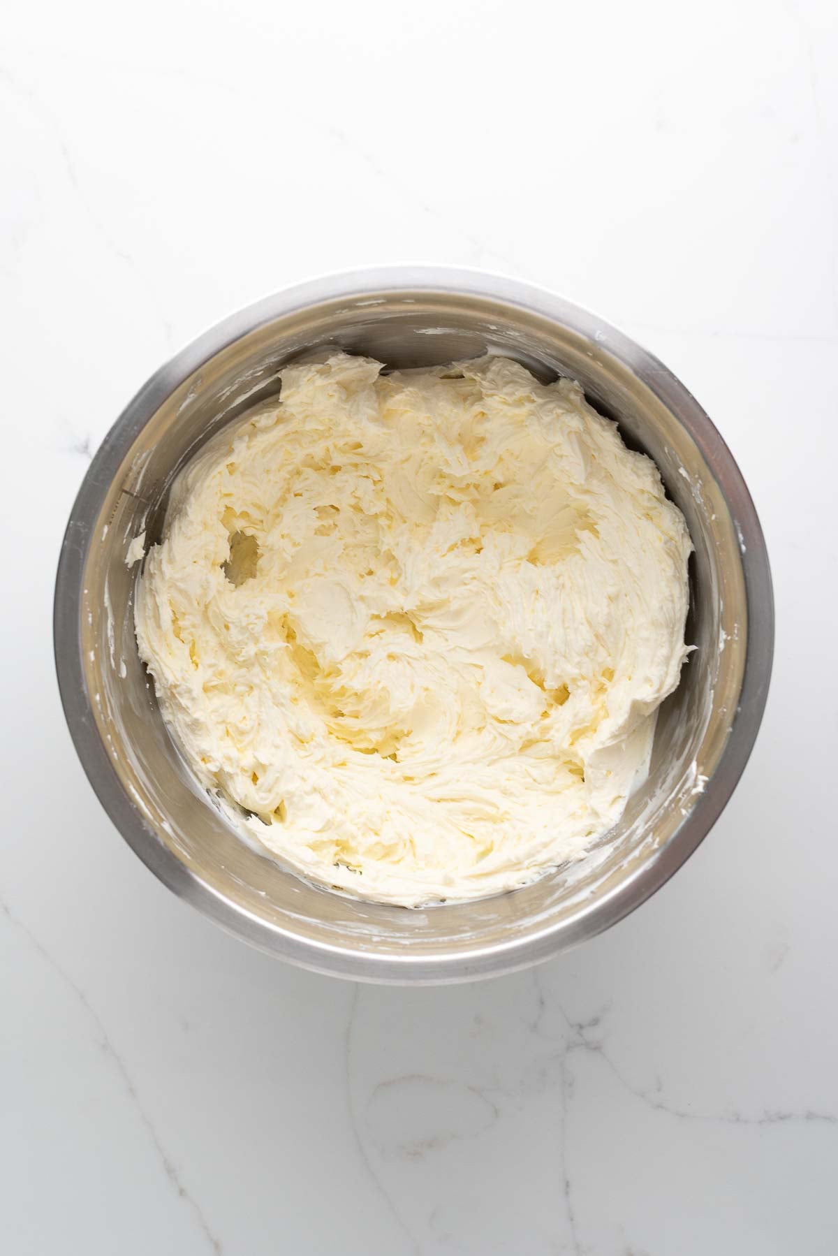 Whipped cream cheese in a stainless steel mixing bowl on a white marbled background.