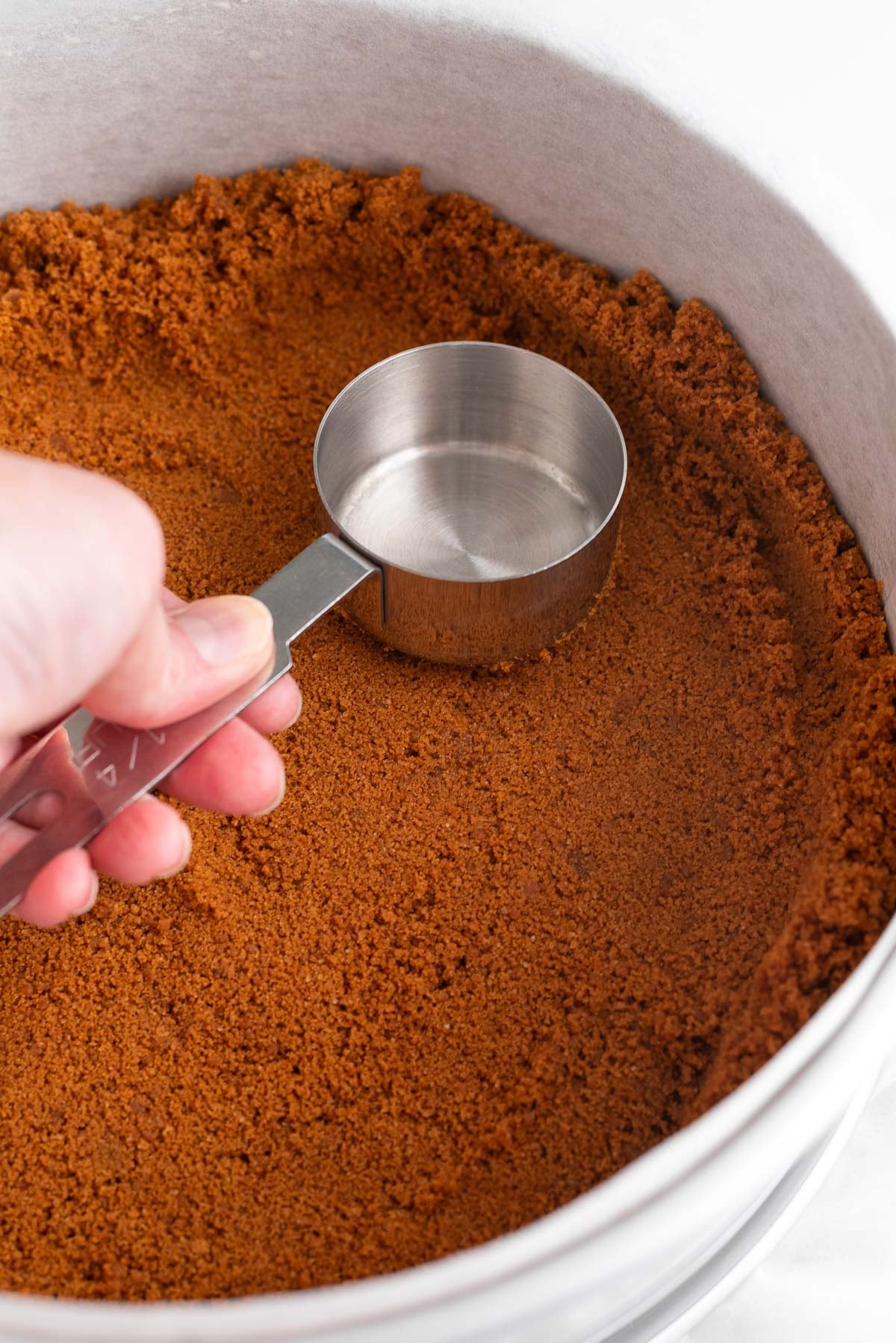 Forming the speculoos cookie crust in a parchment paper lined springform pan using a small metal measuring cup.