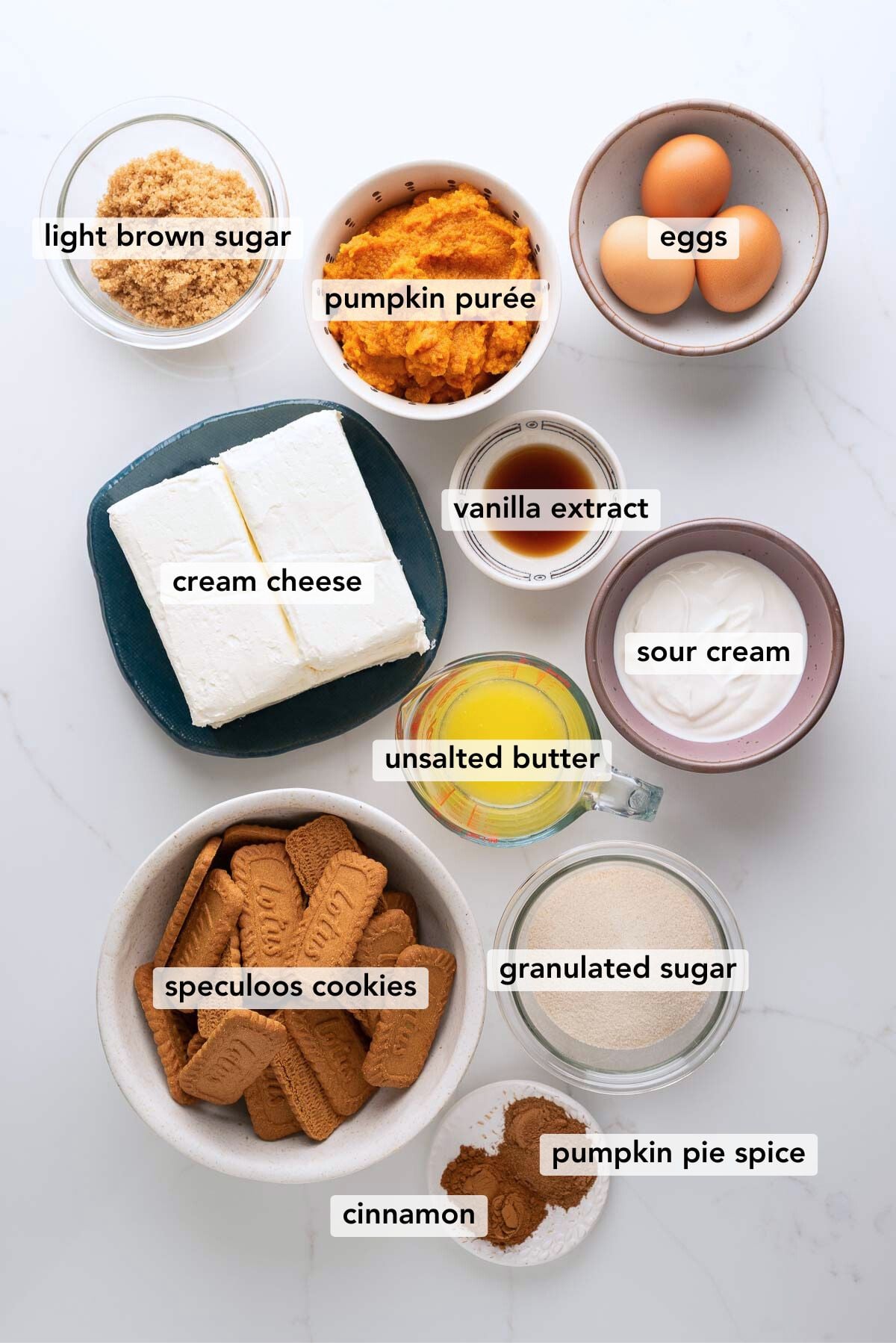 Ingredients in various size bowls labeled as light brown sugar, pumpkin puree, eggs, cream cheese, vanilla extract, unsalted butter, sour cream, speculoos cookies,granulated sugar, cinnamon, and pumpkin pie spice on a white marbled background.