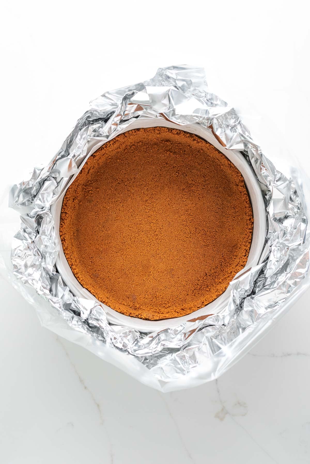 Biscoff cookie crust in a parchment paper lined springform pan wrapped with sheets of aluminum foil and a slow cooker liner.
