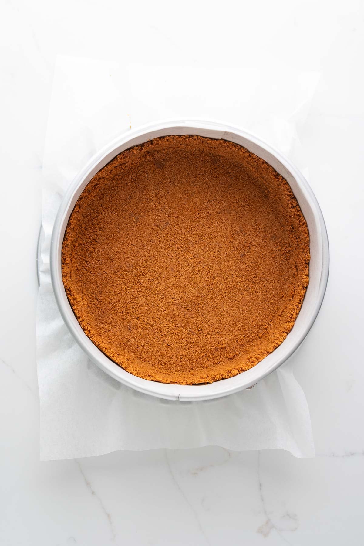 Baked speculoos cookie crust in a parchment paper lined springform pan on a white marbled background.