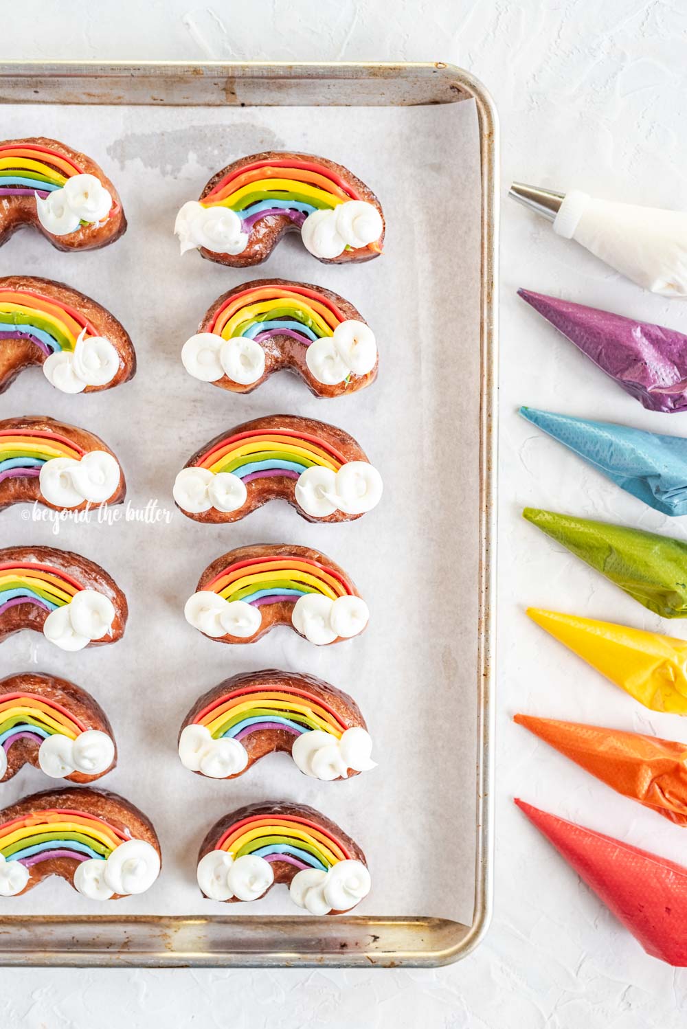 Overhead image of just decorated rainbow donuts on a baking sheet with piping bags filled with buttercream | All Images © Luyiz, LLC