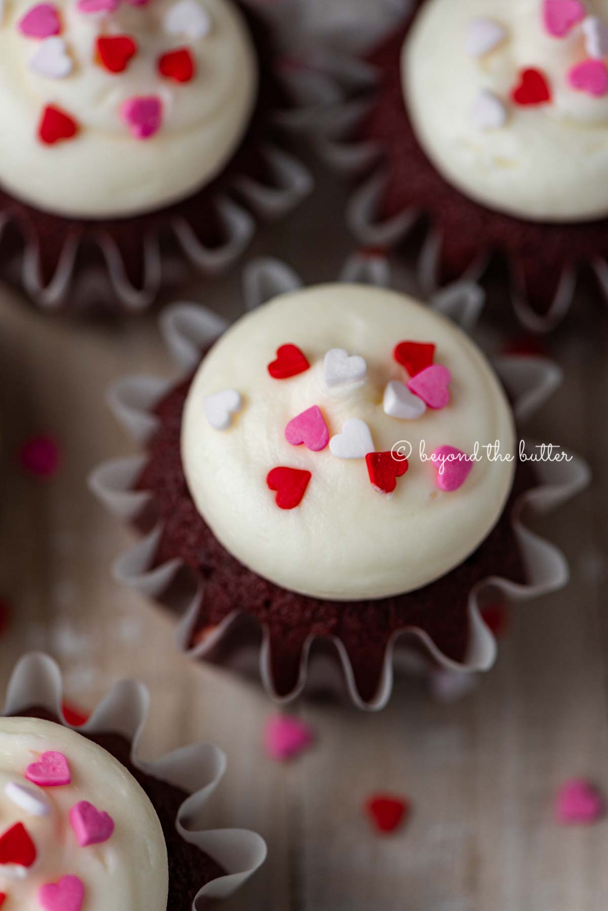 Homemade red velvet cupcakes with cream cheese frosting topped with heart sprinkles on a light wood background | All images © Luyiz®