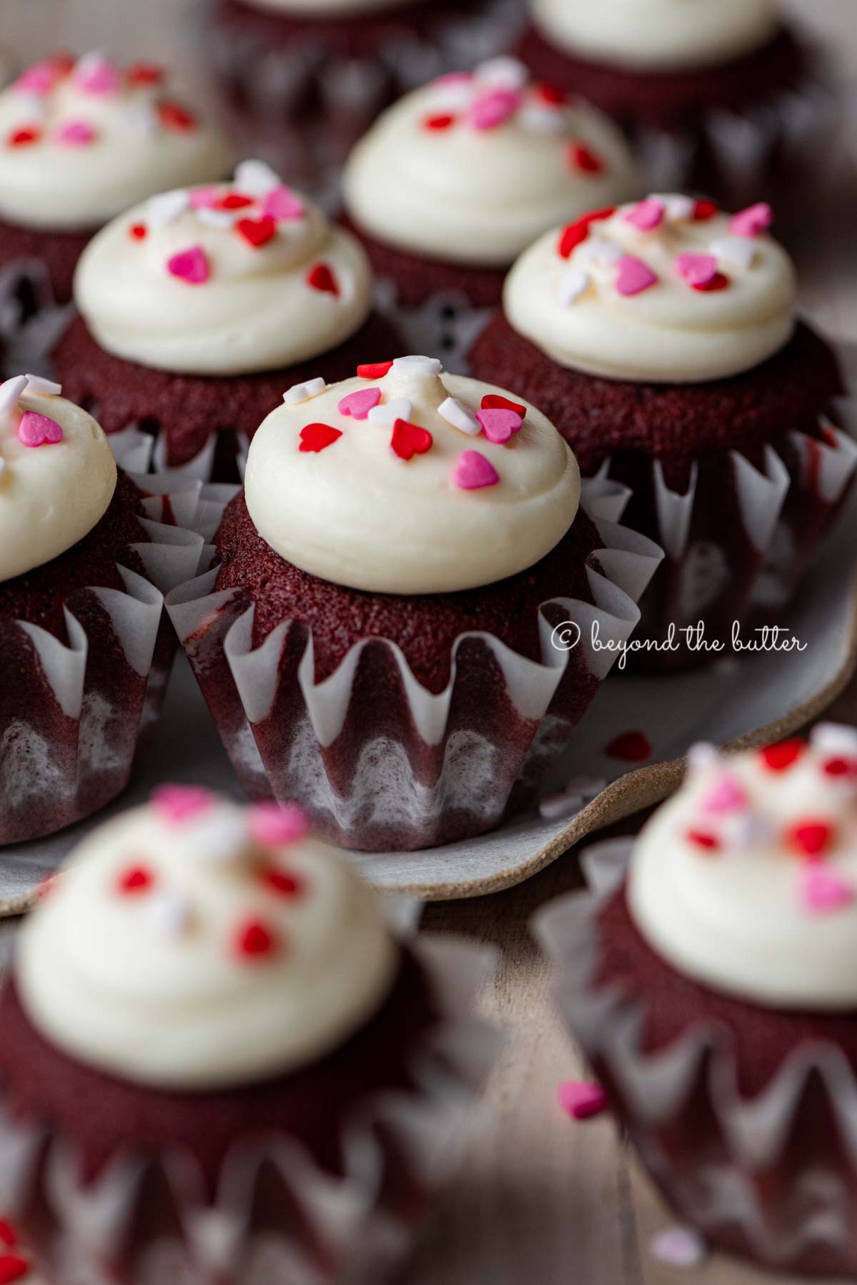 Dessert plate with homemade red velvet cupcakes with cream cheese frosting topped with heart sprinkles on a light wood background | All images © Luyiz®
