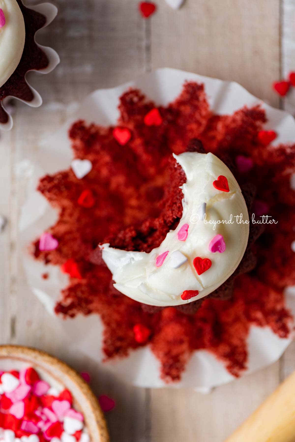 Homemade red velvet cupcakes with one opened and half eaten with a small bowl of Valentine's Day sprinkles on light wood background | All images © Luyiz®