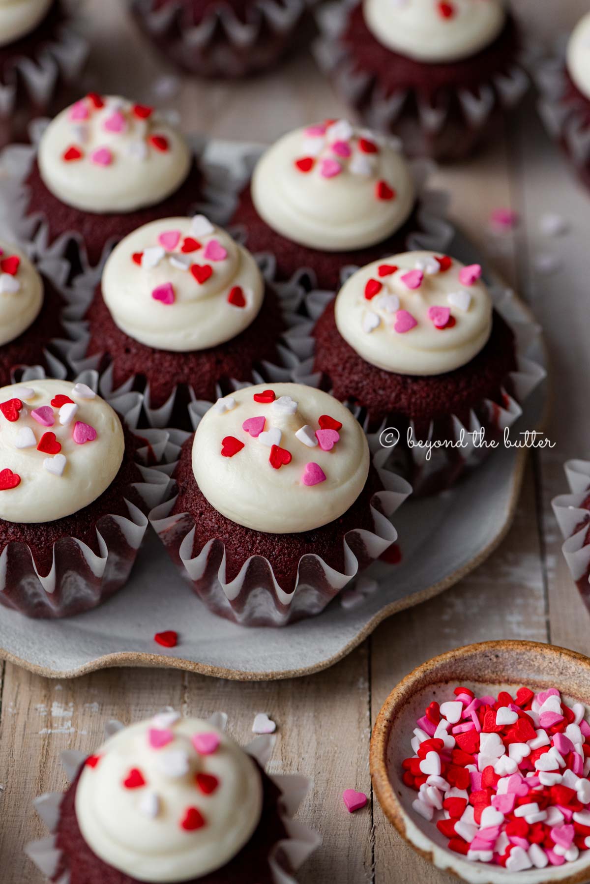 Dessert plate with homemade red velvet cupcakes with cream cheese frosting topped with heart sprinkles on a light wood background | All images © Luyiz®