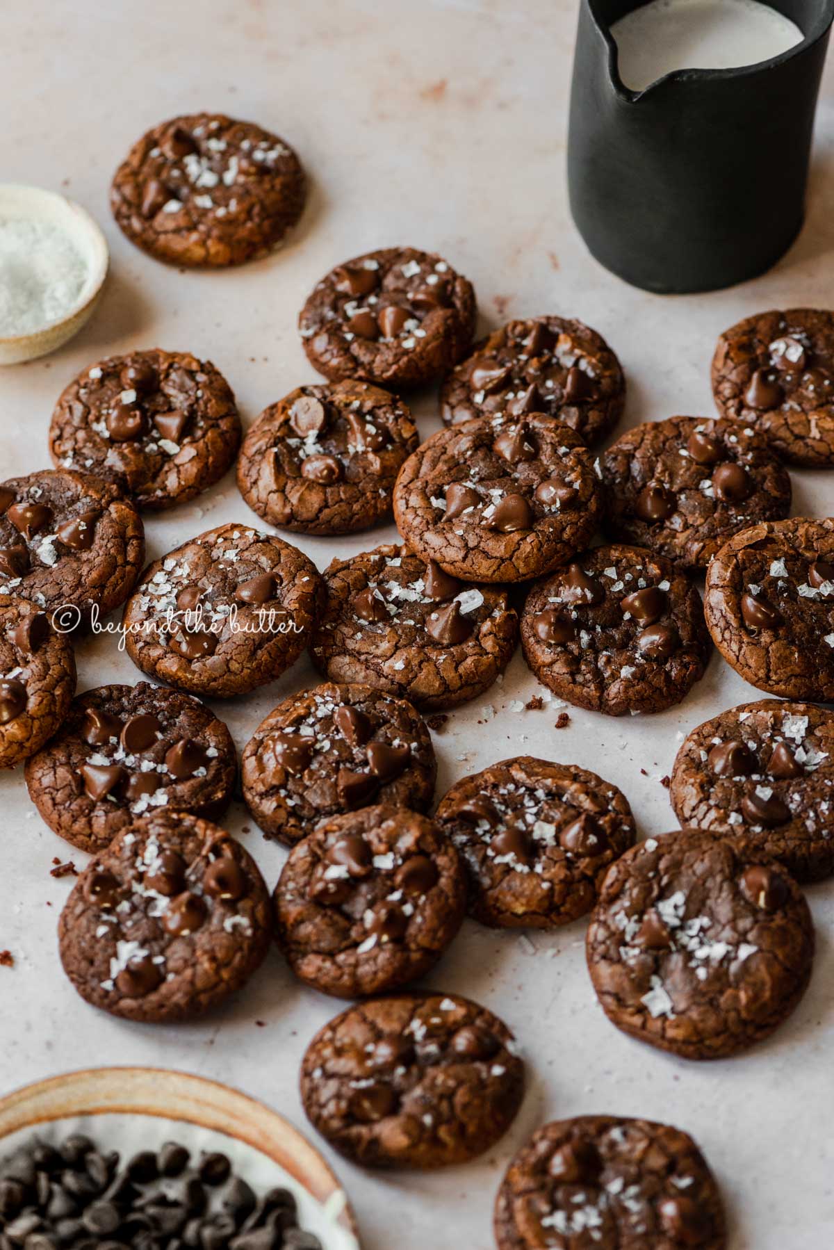 Salted chocolate chip brownie cookies randomly placed on an abstract light brown background with small black pitcher of milk and bowl of sea salt flakes to the side | All Images © Luyiz®