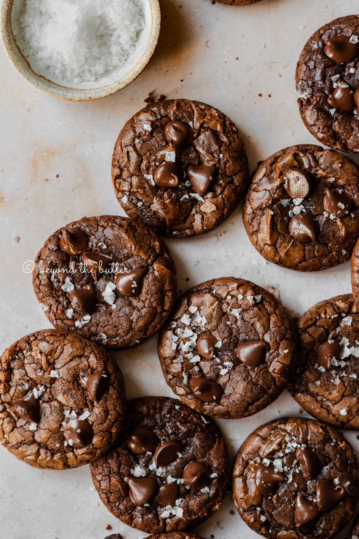 Salted chocolate chip brownie cookies randomly placed on an abstract light brown background with small bowl of sea salt flakes to the side | All Images © Luyiz®