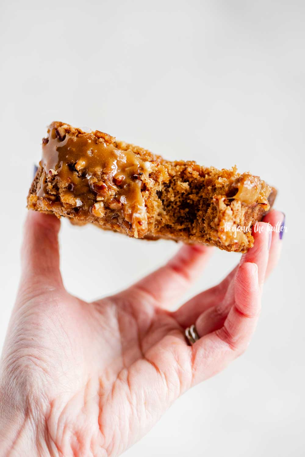 Image of woman's hand hold a slice of salted caramel banana nut bread to show the top with a bite out of it | All Images © Luyiz™