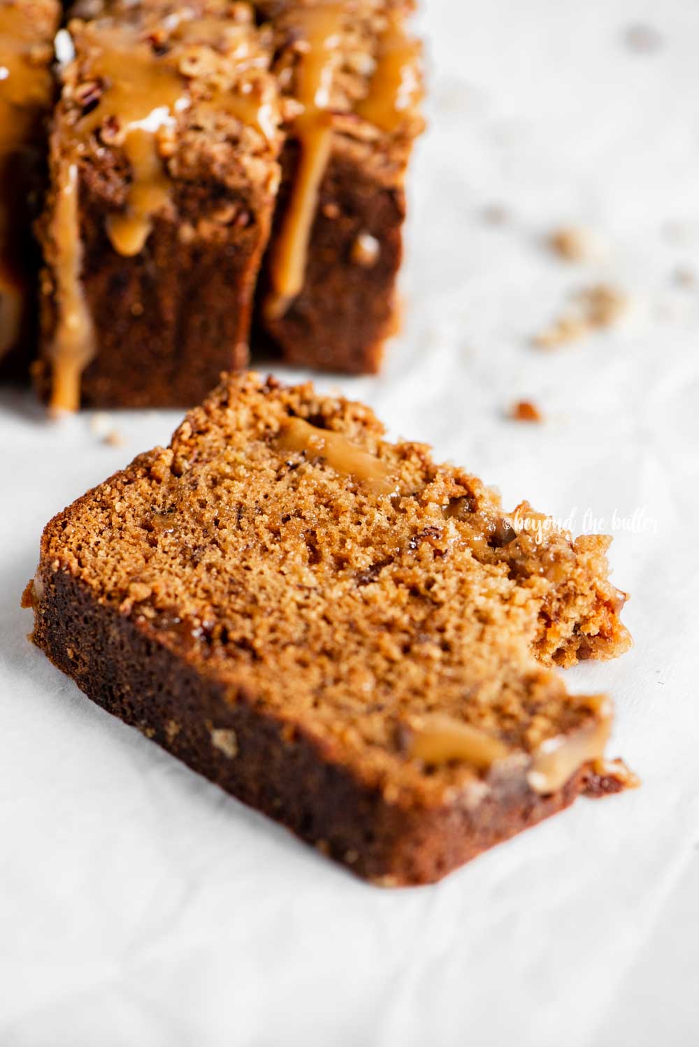 Angled image of slices of salted caramel banana nut bread on parchment paper with a bread knife next to it | All Images © Luyiz™
