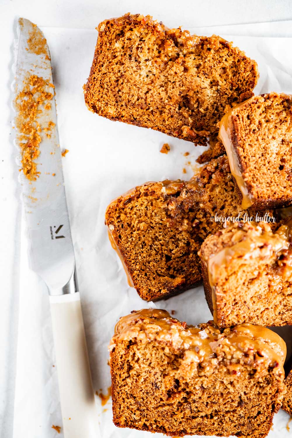 Closeup overhead image of slices of salted caramel banana nut bread on parchment paper with a bread knife next to it | All Images © Luyiz™